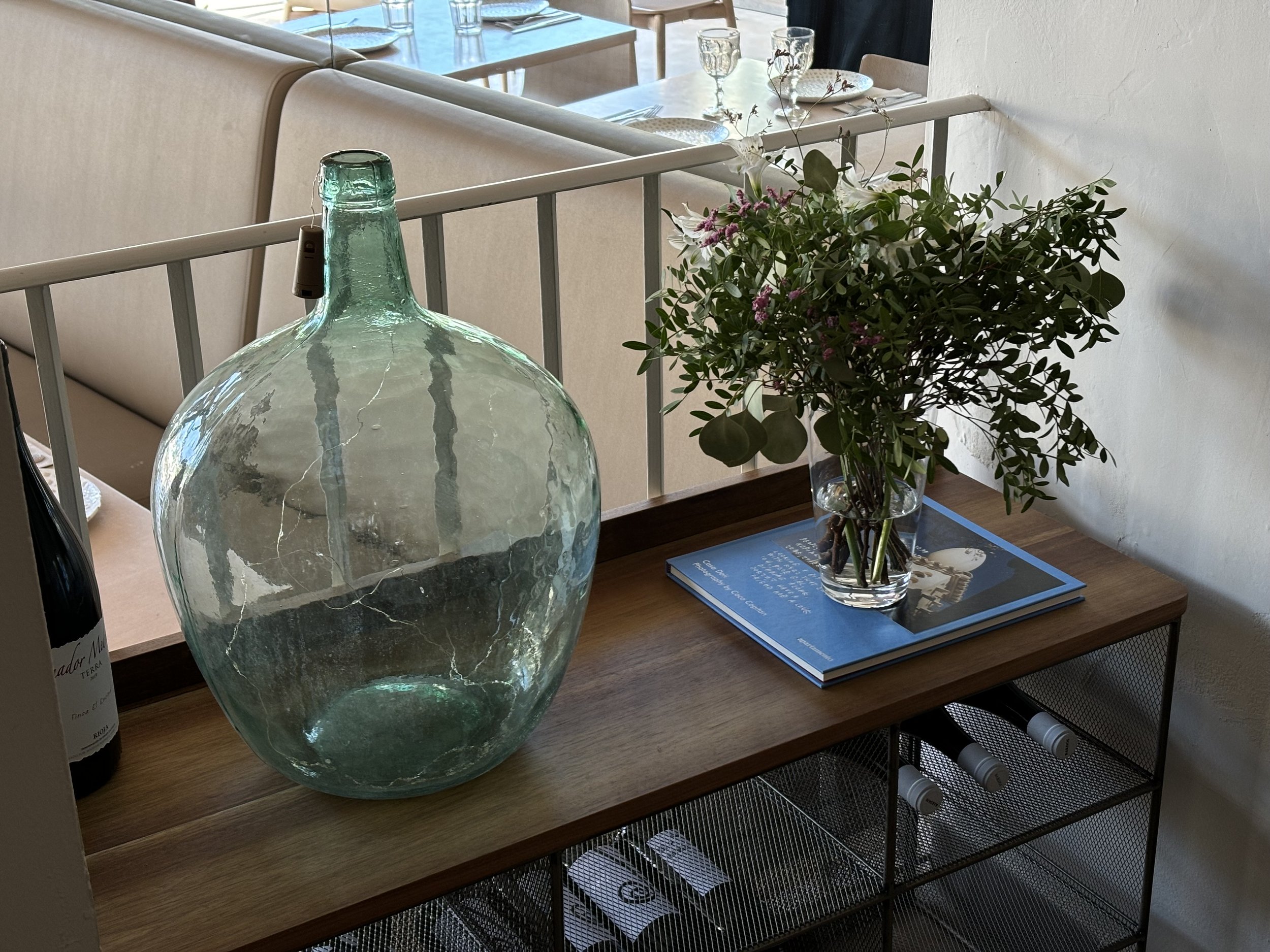 Jarrón grande de vidrio junto a un ramo de flores en una mesa de madera, en un espacio de comedor.