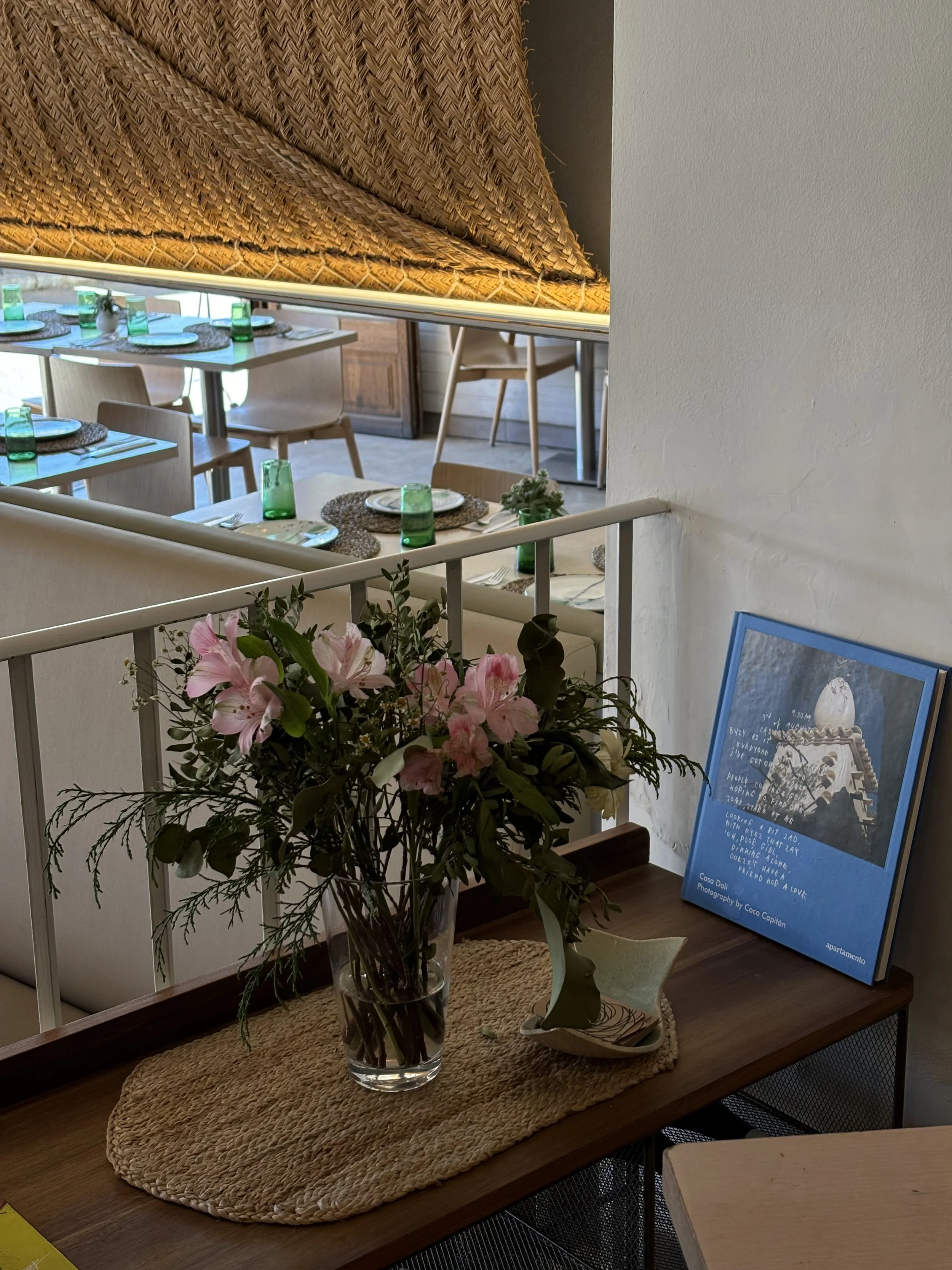 Vista de una sala de restaurante con mesas puestas y un centro de mesa con flores. En primer plano, una mesa de madera con un florero con flores rosadas y un libro de Apartamento, en el fondo, mesas con cristalería y platos, y una alfombra de paja.