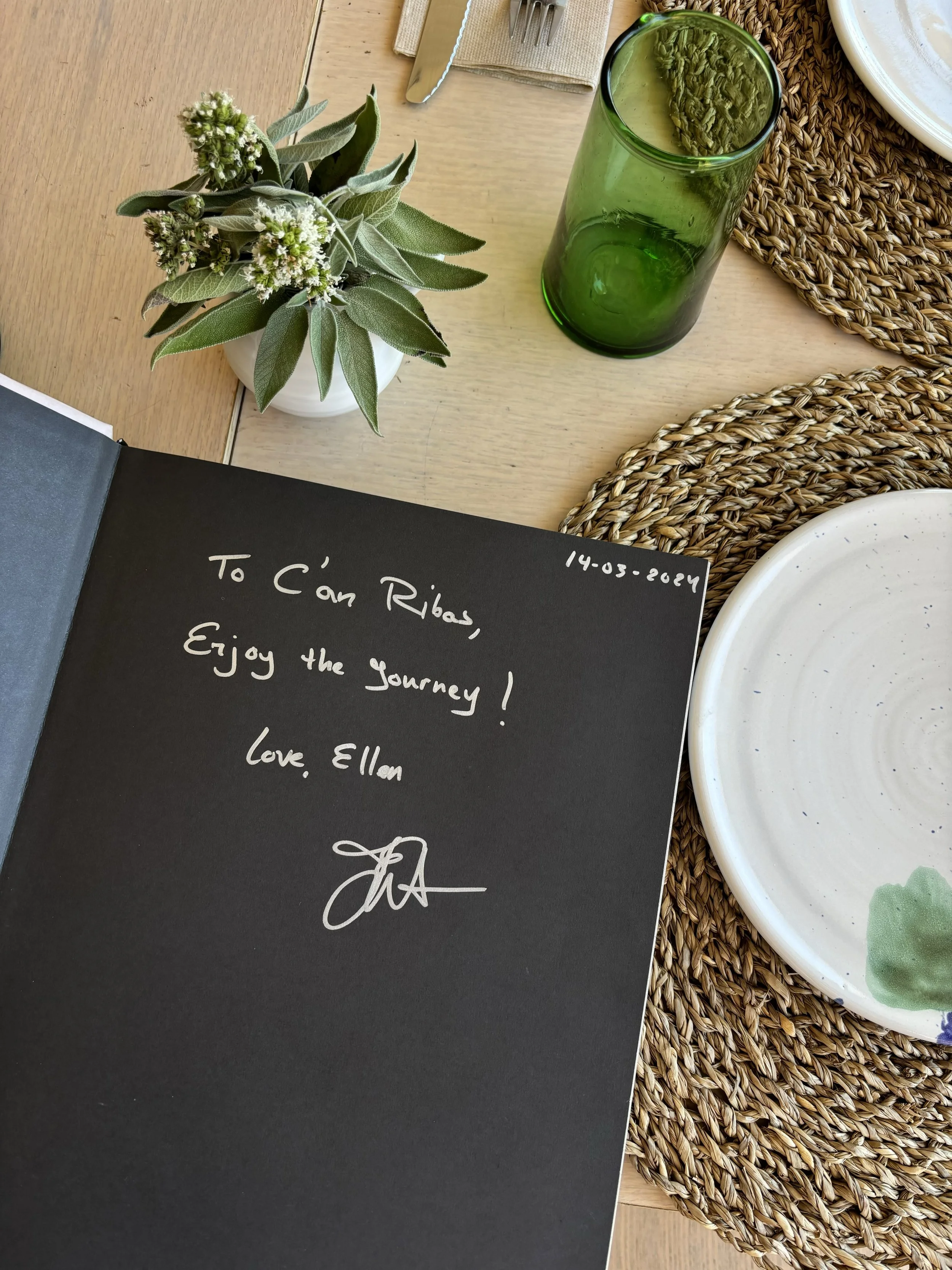 Mesa con una planta, un vaso verde, platos de cerámica y un libro firmado por la autora con mensaje escrito a mano. El libro es sobre Sóller.