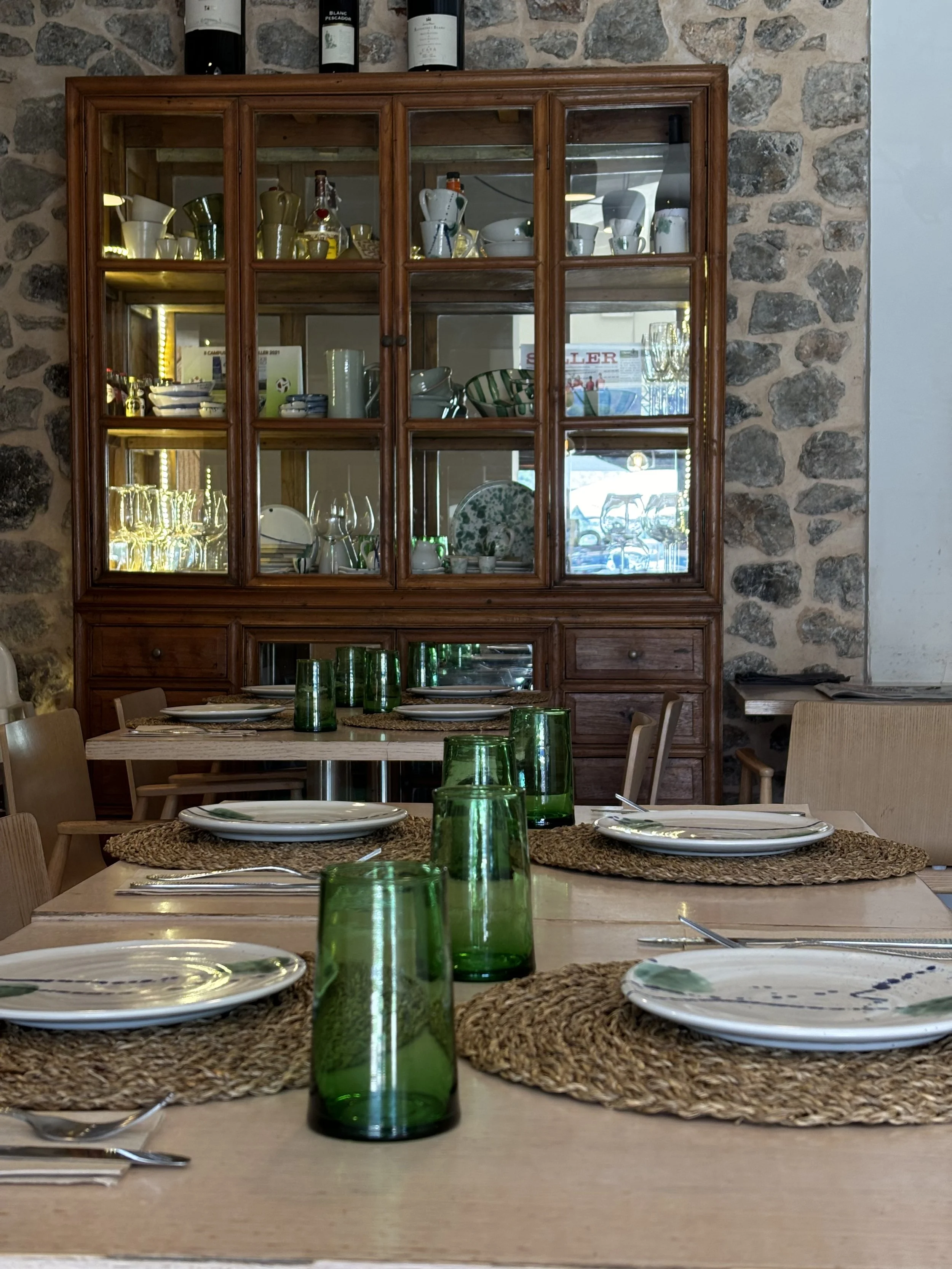 Mesa de comedor con platos, tenedores, cuchillos y vasos verdes en el restaurante Ca'n Ribes, con una vitrina de madera llena de vajilla de Mallorca y cristalería en el fondo.