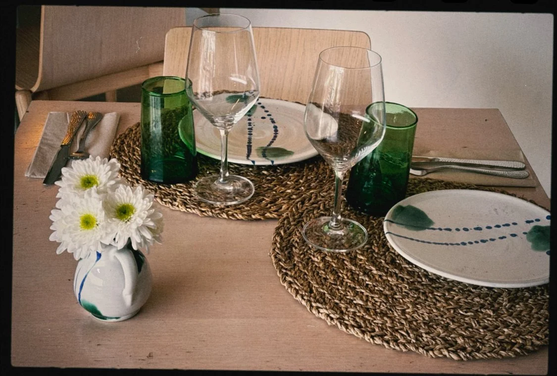 Mesa preparada para una comida con dos platos grandes, dos copas de vino, vasos verdes, cubiertos y un florero con flores blancas.