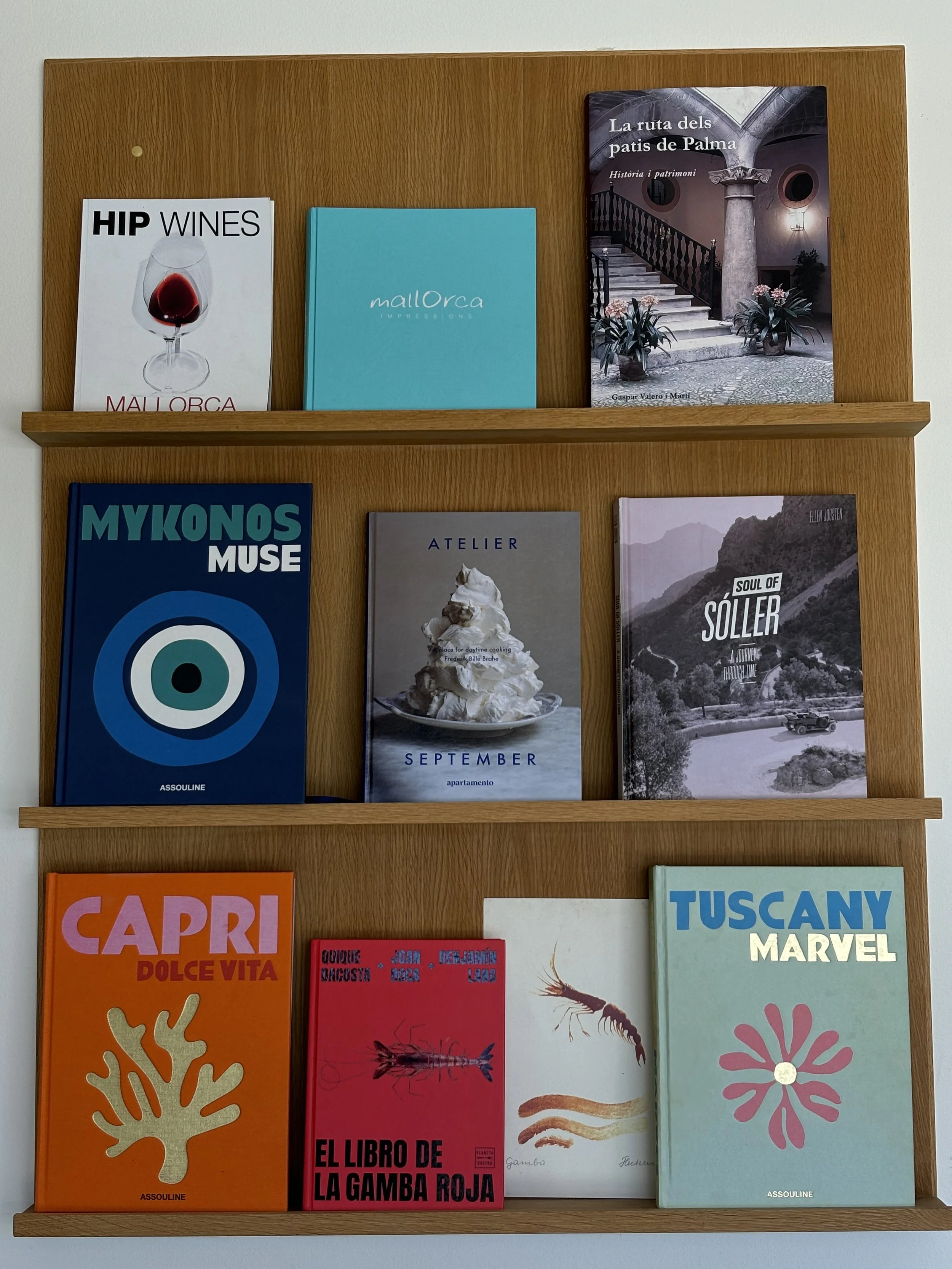 Estantería de madera con libros de diversos temas; en la primera fila hay libros sobre Mallorca y un libro de historia de Palma, en la segunda fila hay libros sobre Mykonos, un taller de cocina, y Sóller, y en la tercera fila hay libros sobre Capri, 