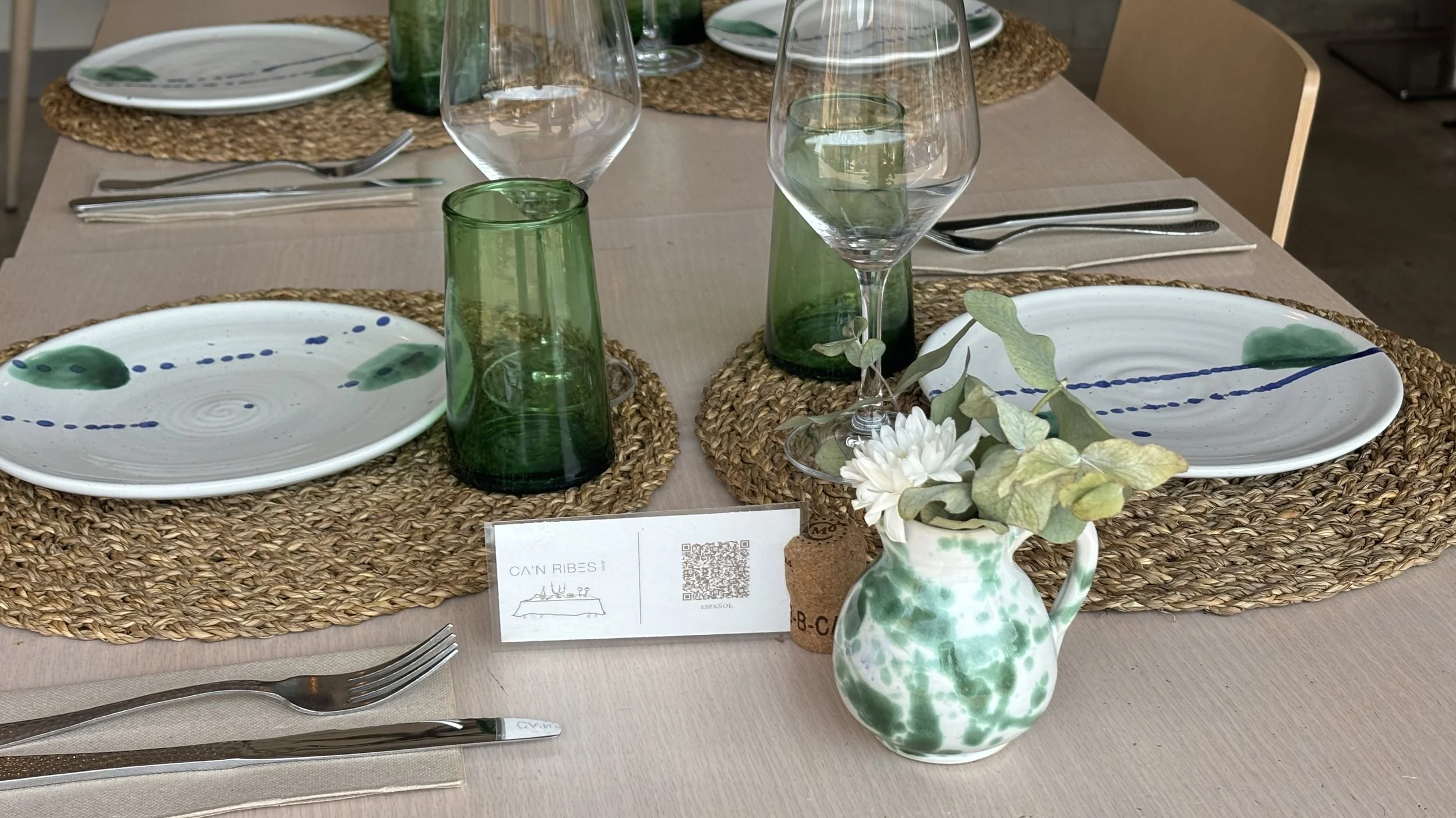 Configuración de mesa con platos decorativos blancos con jarrones verdes y azules con flores dentro, copas de vino y vasos con cristalería verde, centros de mesa con flores blancas y hojas verdes, manteles y platos de cuerda natural.