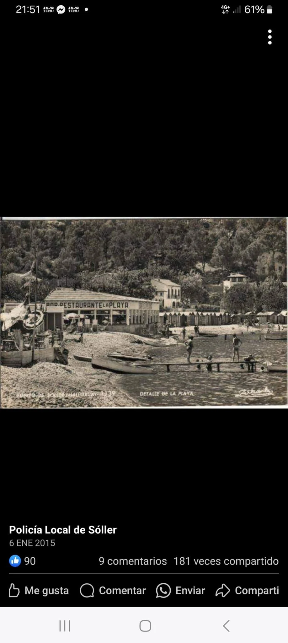 Una vista en blanco y negro de una playa con un restaurante, algunas personas en la orilla y en el agua, y casas en las colinas detrás.