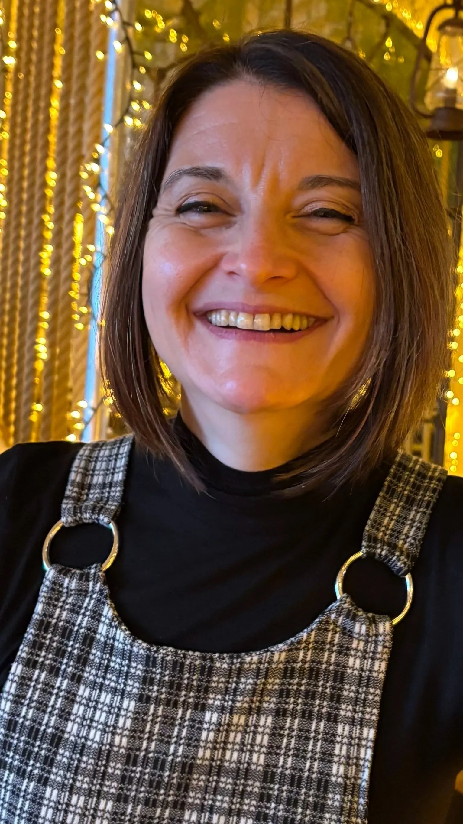 A woman with shoulder-length brown hair smiling in front of a background of warm yellow string lights and wooden decor.