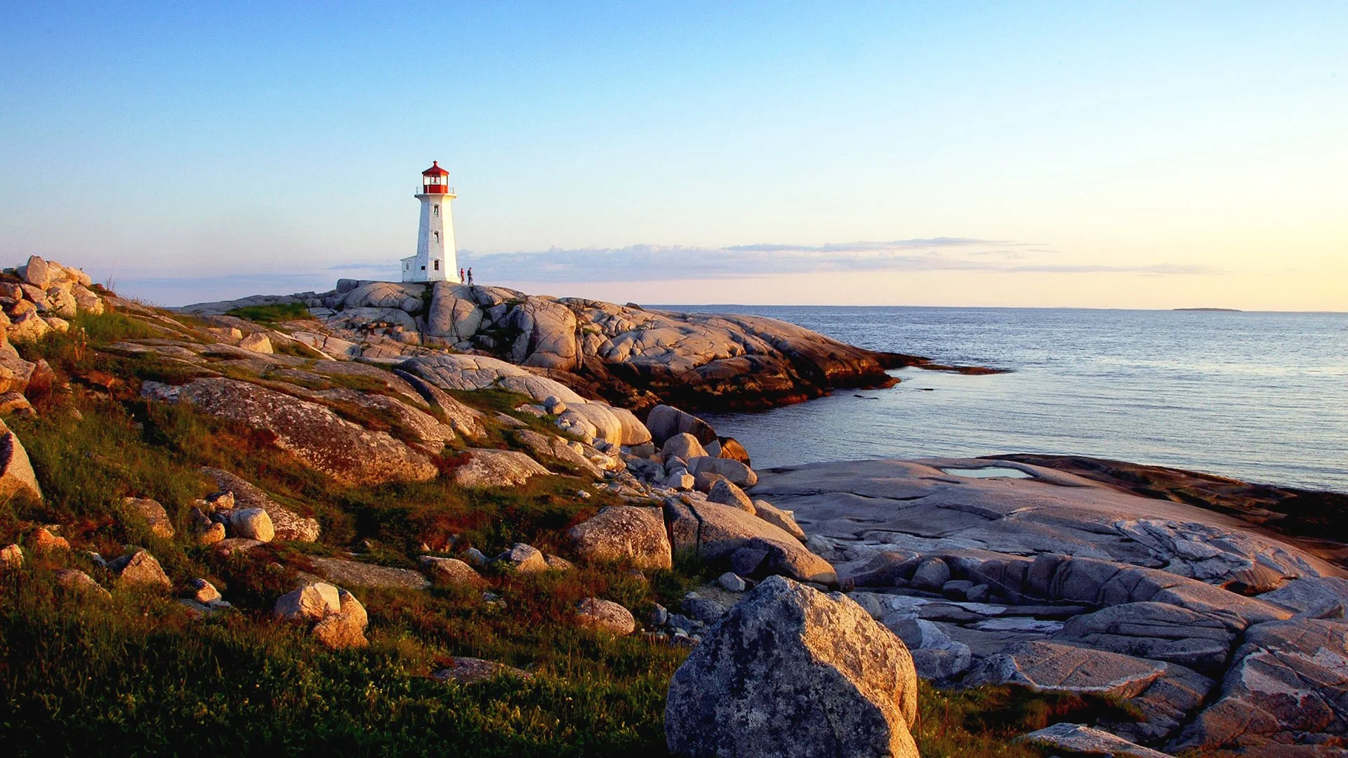 A lighthouse on rocky coast at sunset, overlooking the ocean with a few people nearby.