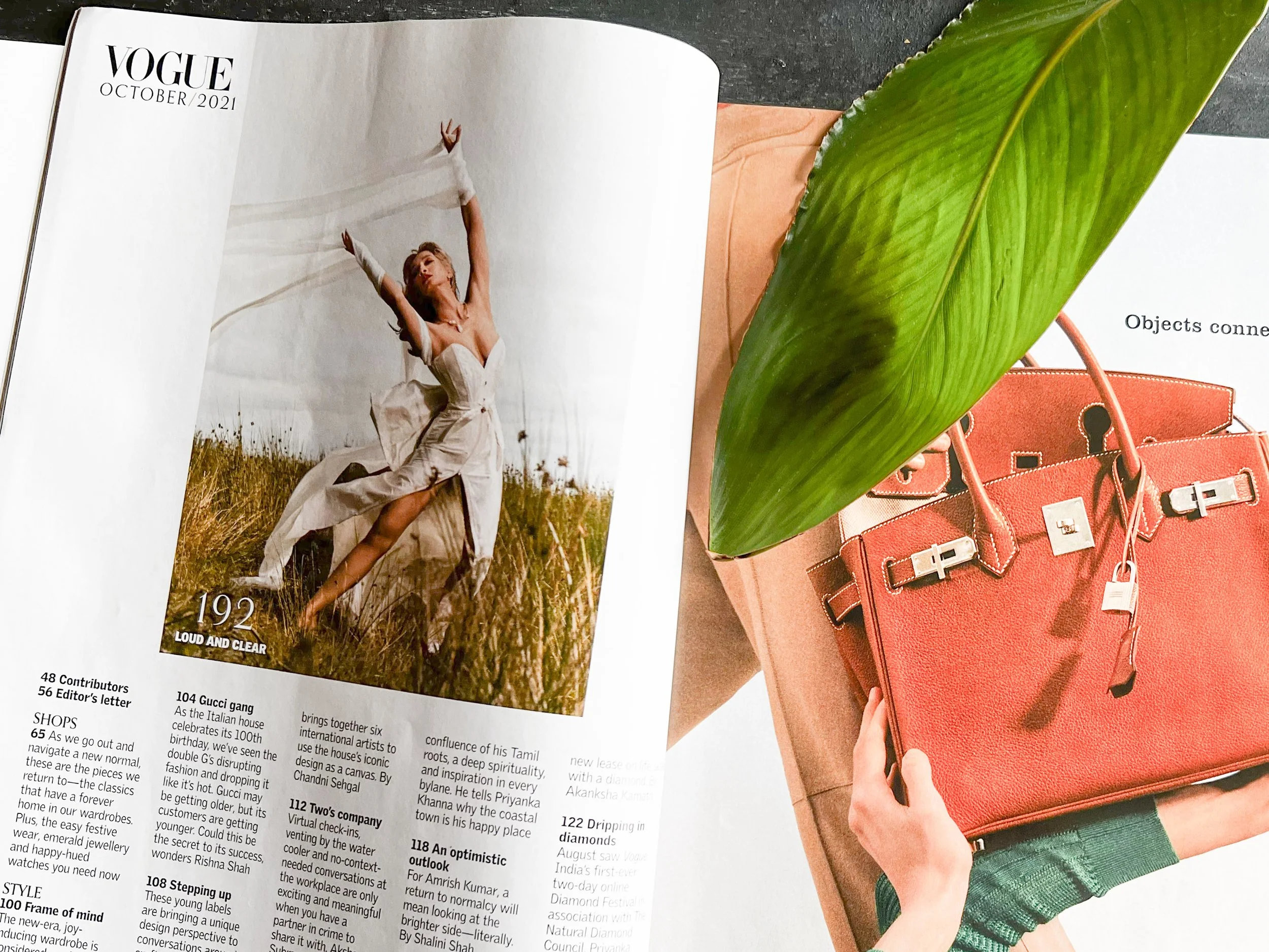 Open Vogue magazine featuring a woman dancing outdoors in a white dress, next to a green leaf and a coral-colored handbag on a pink and black background.
