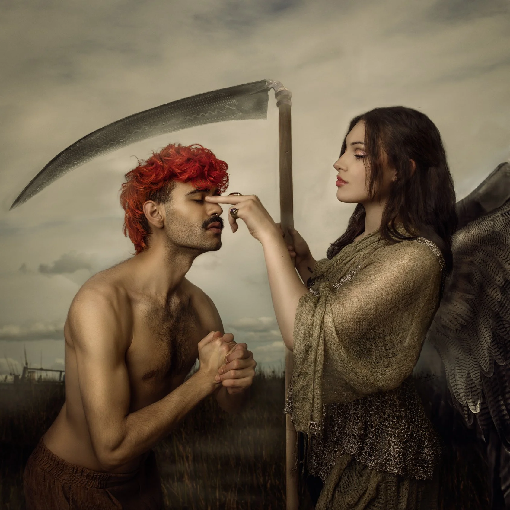 A shirtless man with red curly hair holding his hands together and a woman with dark hair, dressed in earthy tones, gently touching his nose with her finger. The woman has fairy-like wings and is holding a scythe. Photography by Karolina Skorek