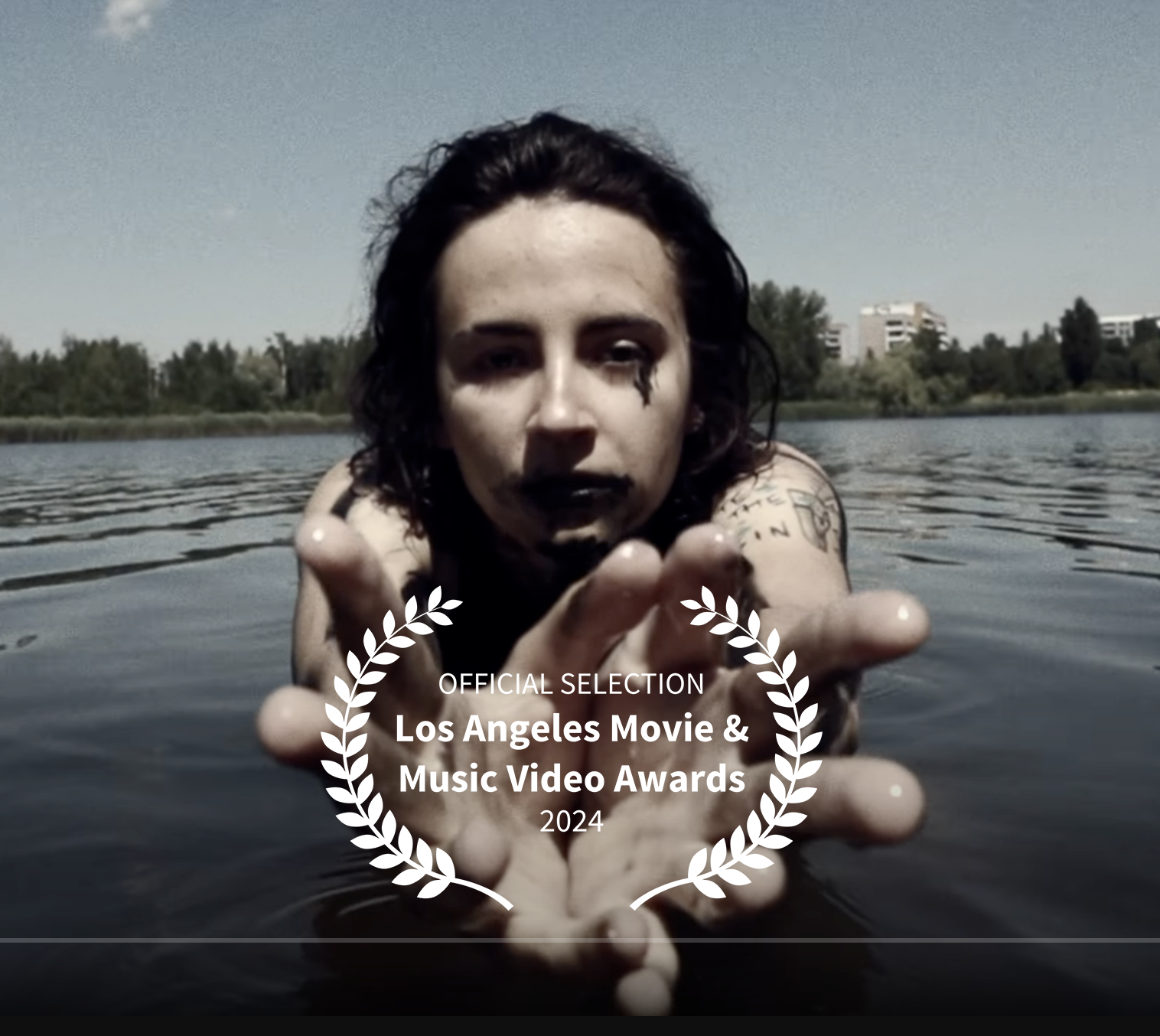 A woman with dark hair and tattoos on her arm reaches out toward the camera while emerging from a body of water, with trees and buildings visible in the background, and an award overlay indicating her film's selection at the Los Angeles Movie & Music