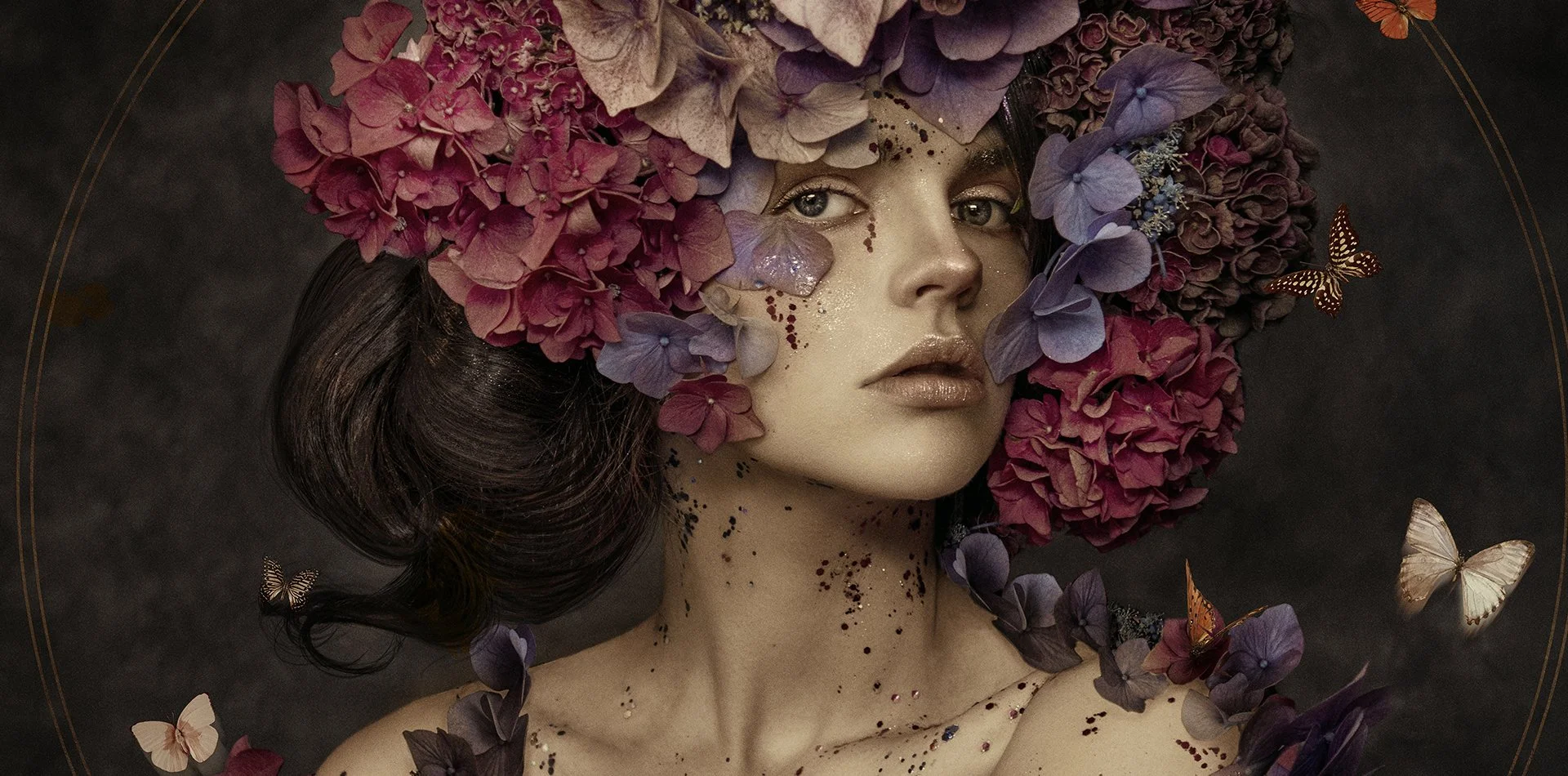 A woman with dark hair and fair skin is adorned with a large headdress made of pink and purple hydrangeas. She has artistic makeup with glitter and small sequins on her face and body, and is surrounded by floating butterflies in tones of white, orang