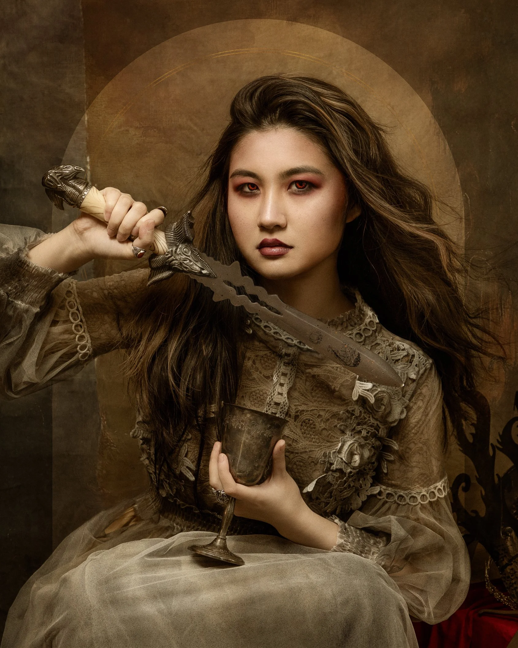 A woman with long brown hair and red eyeshadow seated at a table with a textured beige tablecloth, holding a silver goblet and a large ornate knife, dressed in vintage beige lace clothing with ruffled sleeves, in a dark, richly decorated room with a 