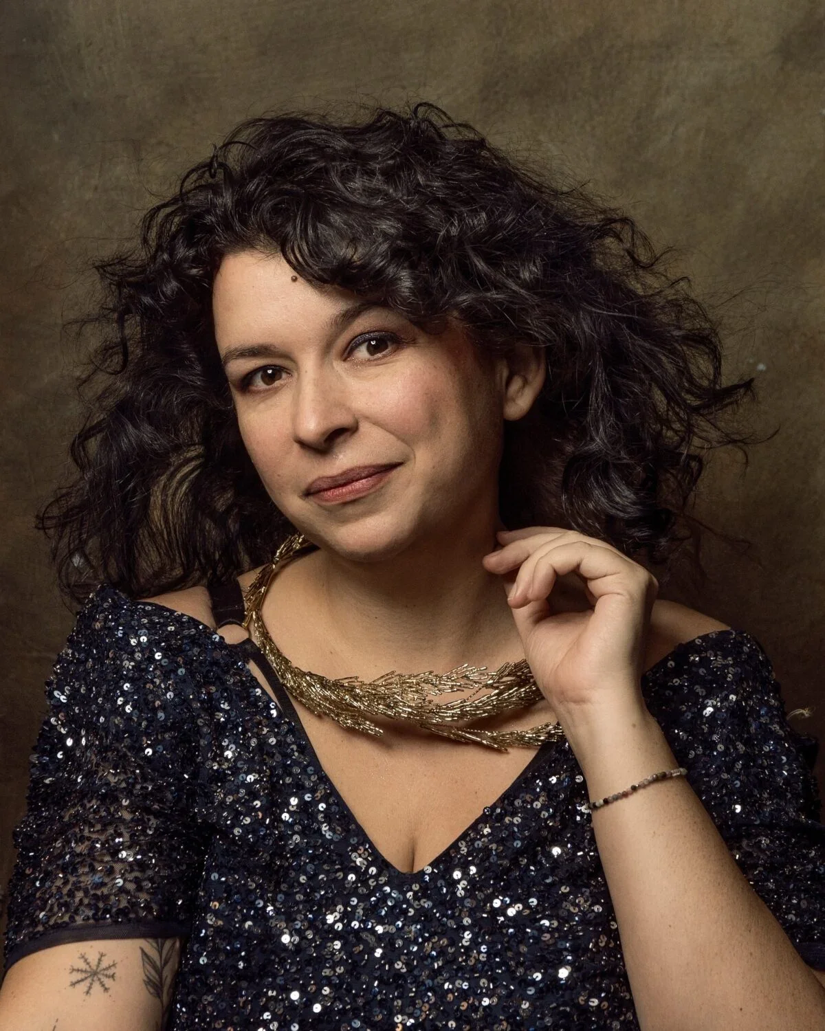 Portrait of a Karolina Skorek - woman with dark, curly hair wearing a sparkly sequin top, layered gold necklace, and a bracelet, with a brown textured background.