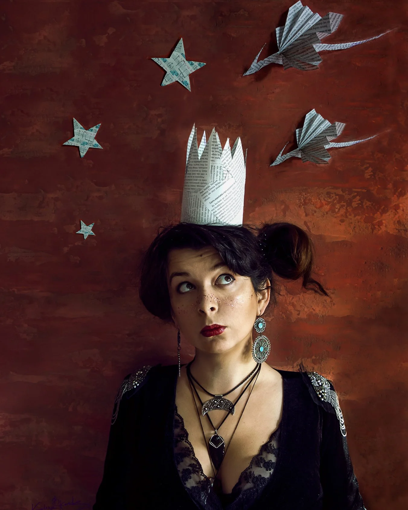 A woman with dark hair and fair skin, wearing large turquoise earrings and layered necklaces, looks upward with a thoughtful expression. She has a makeshift paper crown on her head and three paper stars and two paper dragons on the warm-toned, textured background.