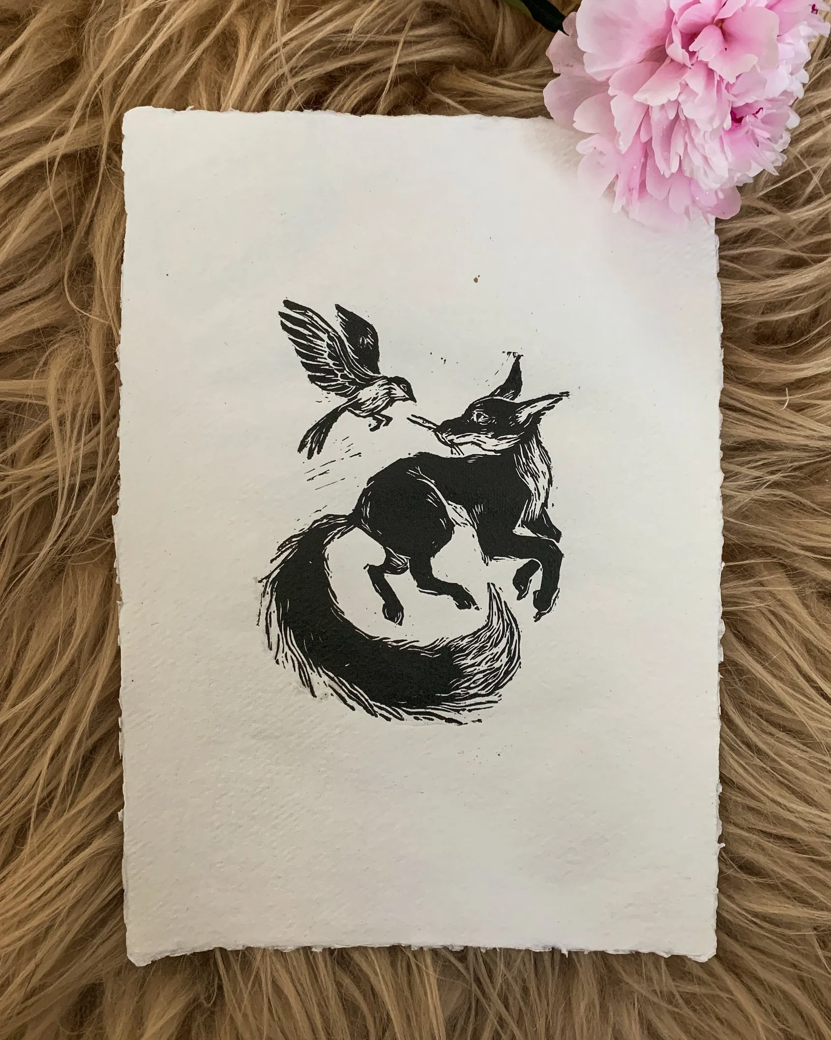 A black and white illustration of a fox with a bird sitting on its nose, on a white textured paper, placed on a furry surface with pink flowers in the corner. by Karolina Skorek