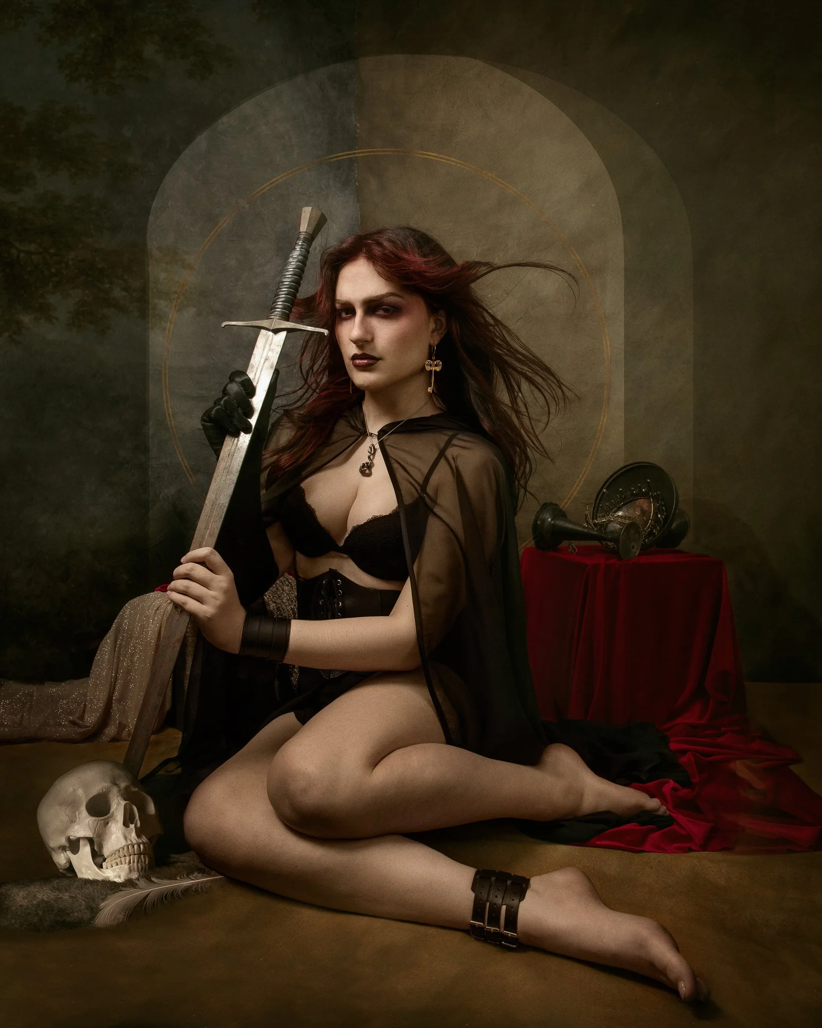 A woman with red hair and dark makeup sitting on the floor with her legs crossed, holding a sword with a skull and a feather nearby, a skull and vintage microphone on a red draped table behind her, and a textured wall with geometric designs in the ba