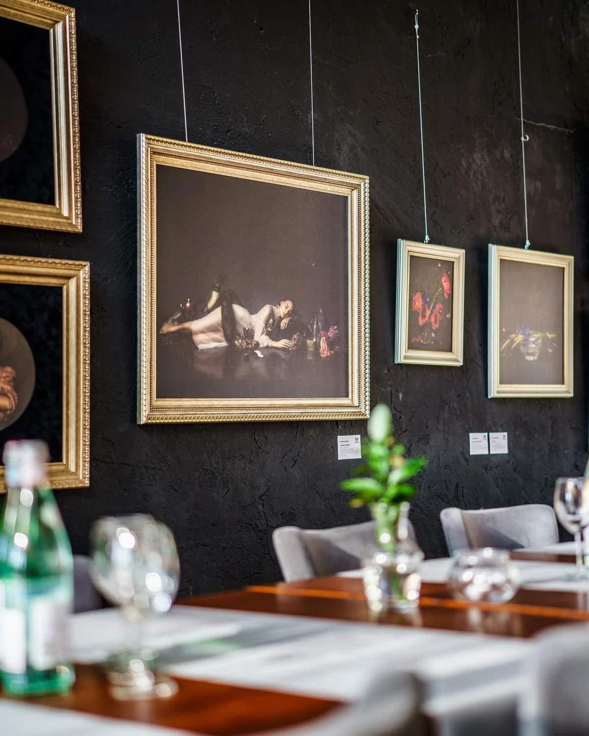 Gallery wall with framed paintings made by Karolina Skorek at a restaurant, with a table set with water bottles, glasses, and a flower in the foreground.