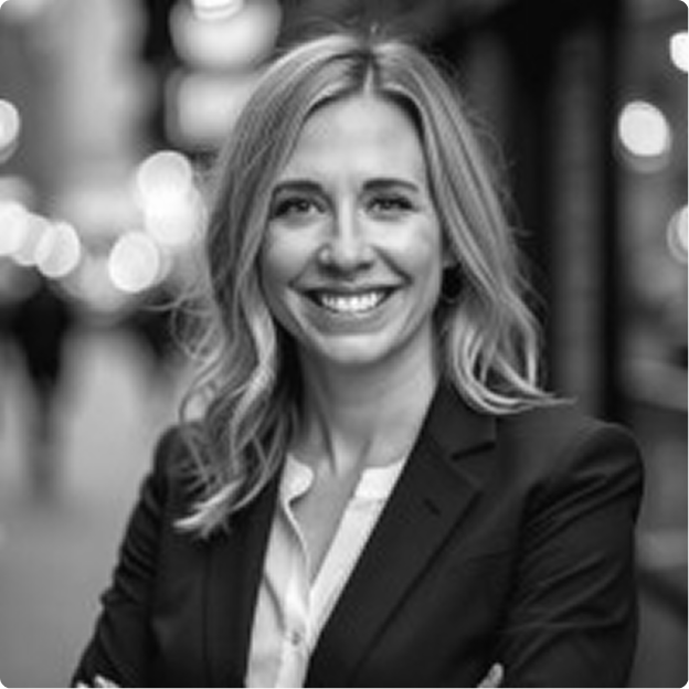 A woman with wavy blonde hair smiling, wearing a blazer and white blouse, standing outdoors with blurred city lights in the background.