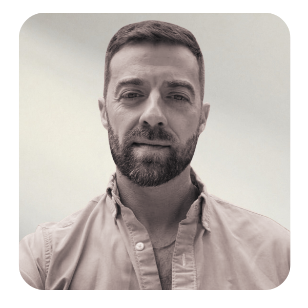 A man with short hair and a beard wearing a beige button-up shirt, looking directly at the camera with a neutral expression.
