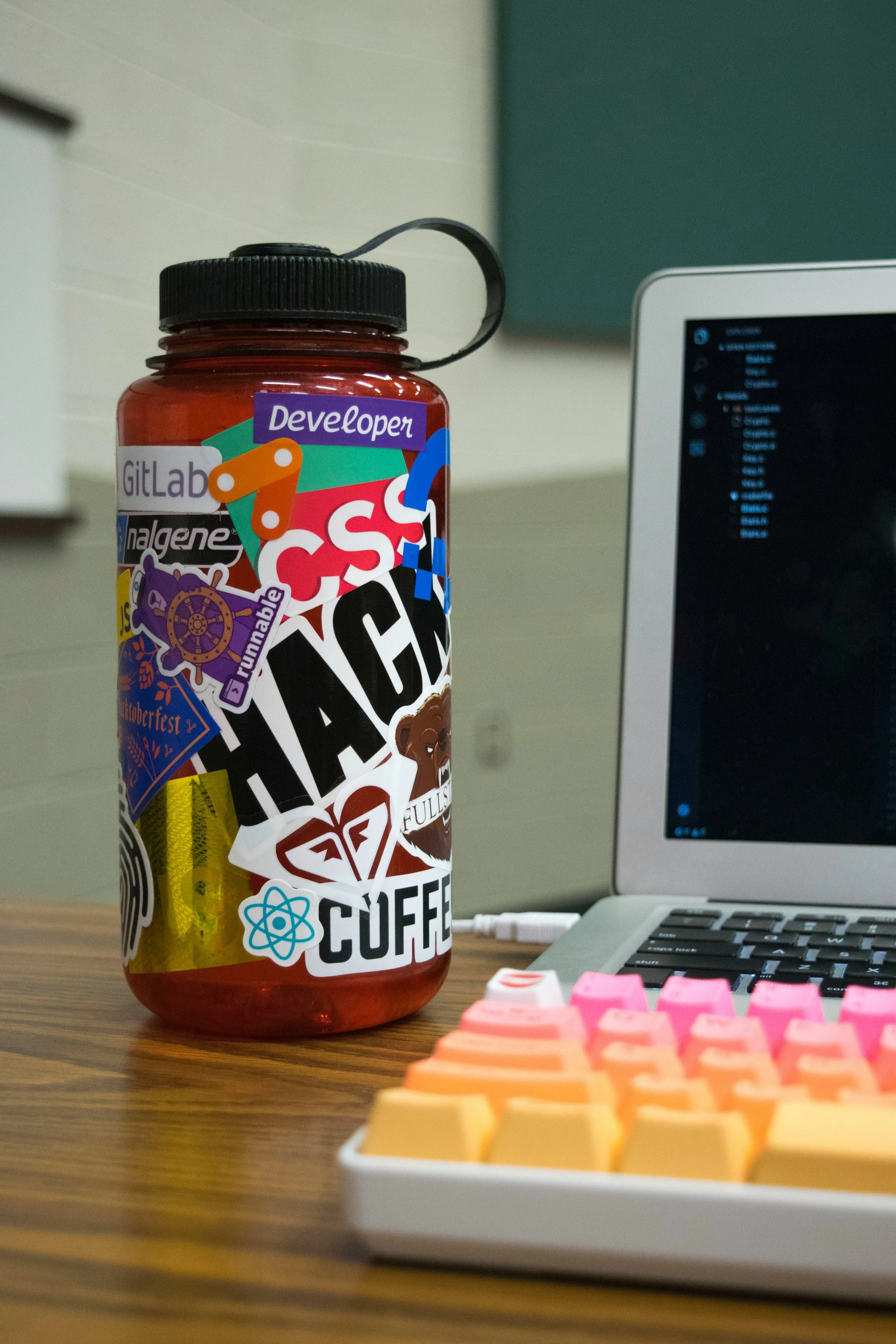 A red water bottle with multiple stickers, including college-related, tech, and novelty designs, on a wooden desk with a laptop and a mechanical keyboard in a home or office setting.