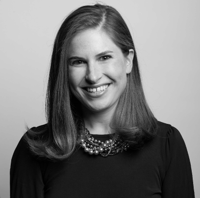 A smiling woman with shoulder-length hair wearing a dark top and a multi-strand pearl necklace, posed against a plain background.