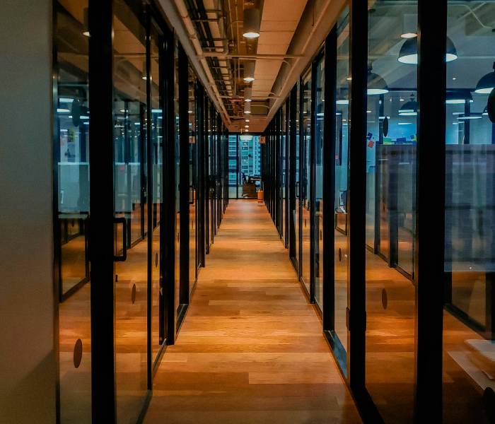Long hallway with glass-walled office spaces on either side, wood flooring, and ceiling lights, in a modern office building.
