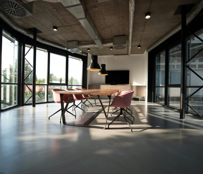 Modern conference room with large windows, a wooden table, pink chairs, pendant lights, a wall-mounted TV, and cityscape view outside.