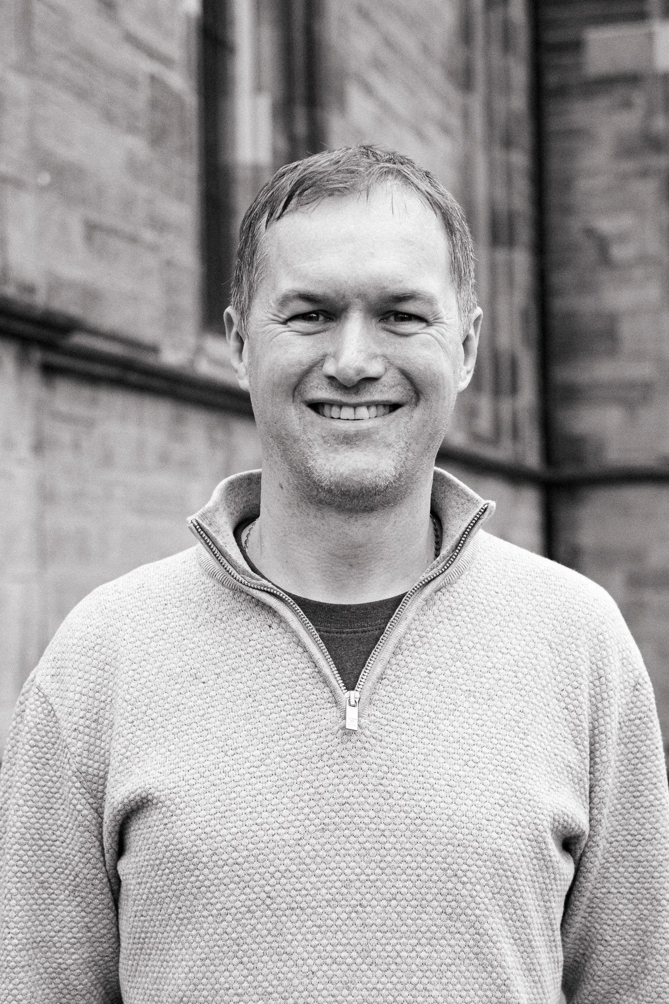 A smiling man wearing a zip-up sweater outside in front of a brick and wooden building, in a black and white photo.