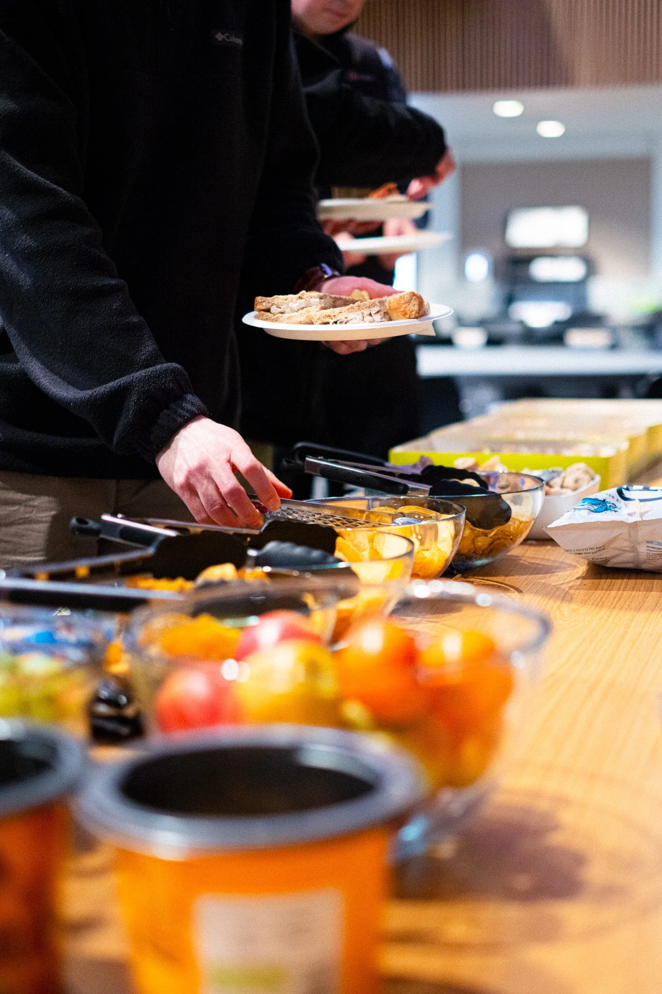 People serving themselves food at a buffet table with various dishes and bowls of fruit.