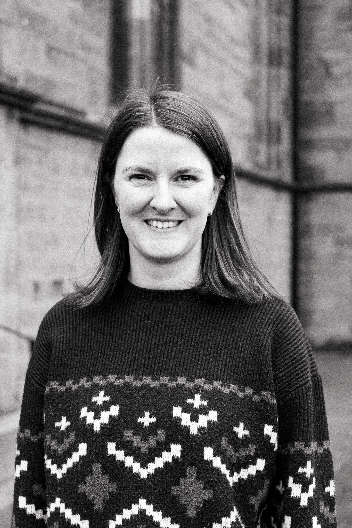 Black and white portrait of a smiling woman with shoulder-length hair, wearing a sweater with a Christmas pattern, standing outdoors in front of a brick wall.