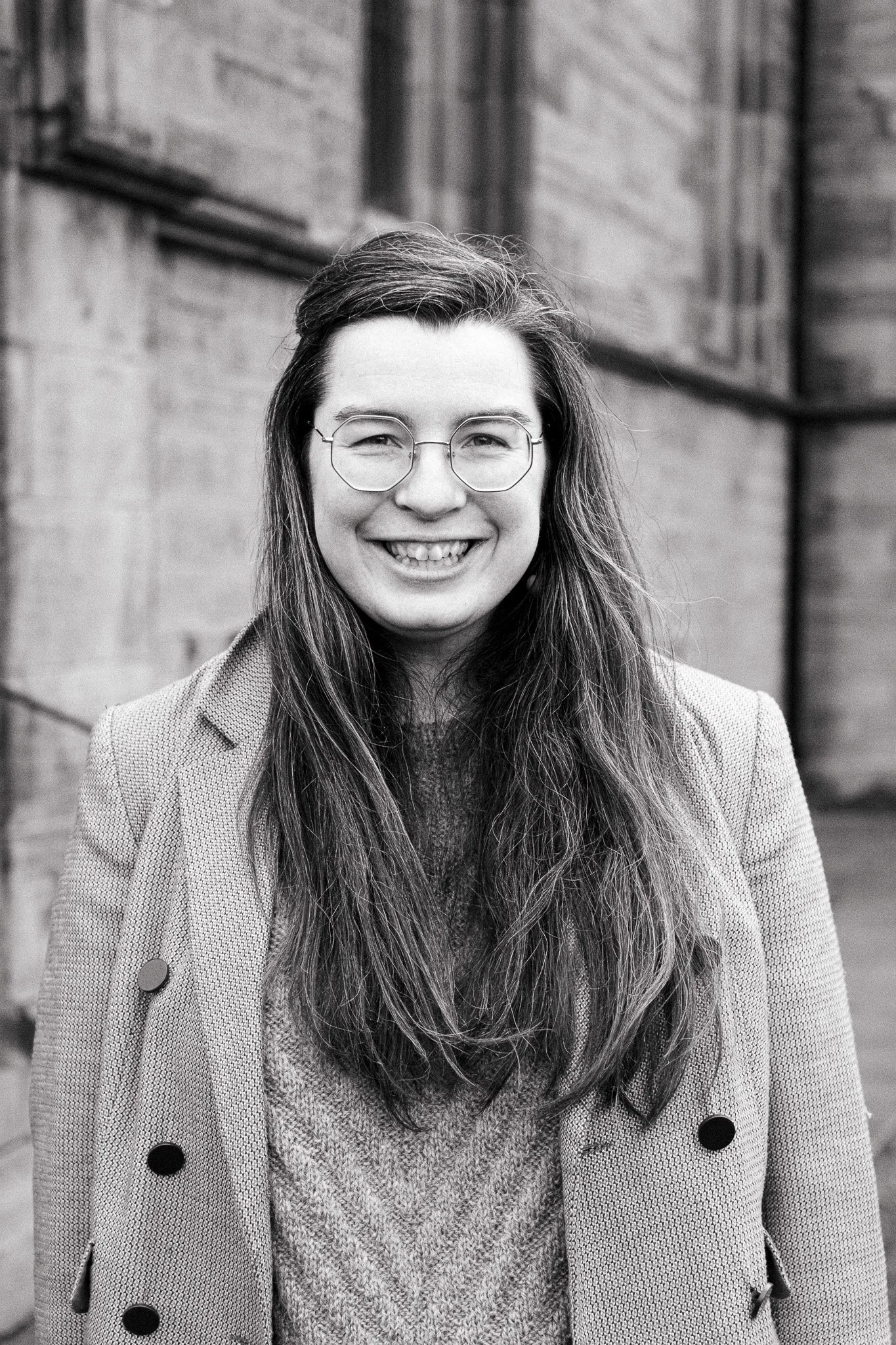 A smiling woman with long hair wearing glasses, a coat, and a sweater, standing outdoors near a brick building.