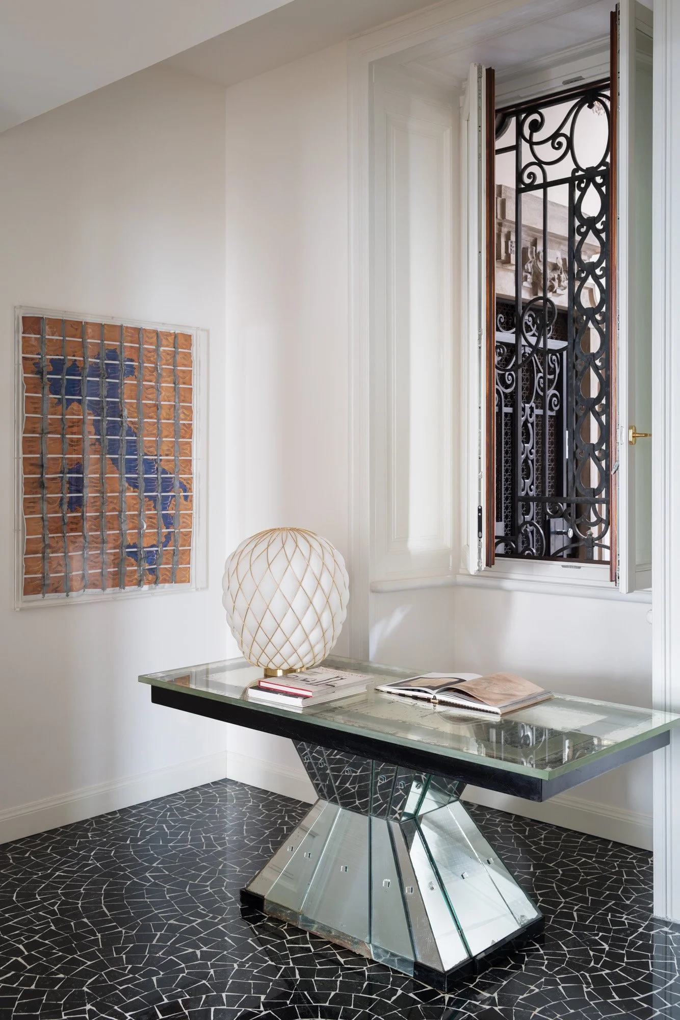 Interno di un appartamento con pareti bianche, il pavimento nero con motivo geometrico, una finestra con grate in ferro battuto e una scrivania con lampada di design e riviste