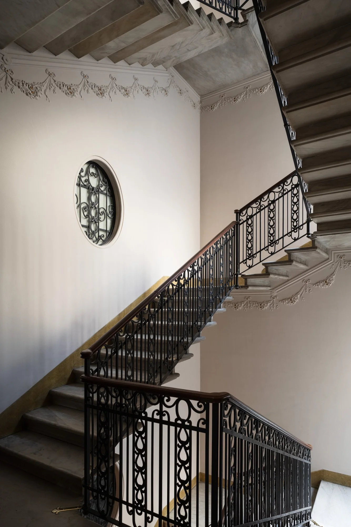 Scalinata con ringhiera in ferro battuto e elementi decorativi in marmo, all'interno di un edificio con pareti bianche e ornamenti decorative.