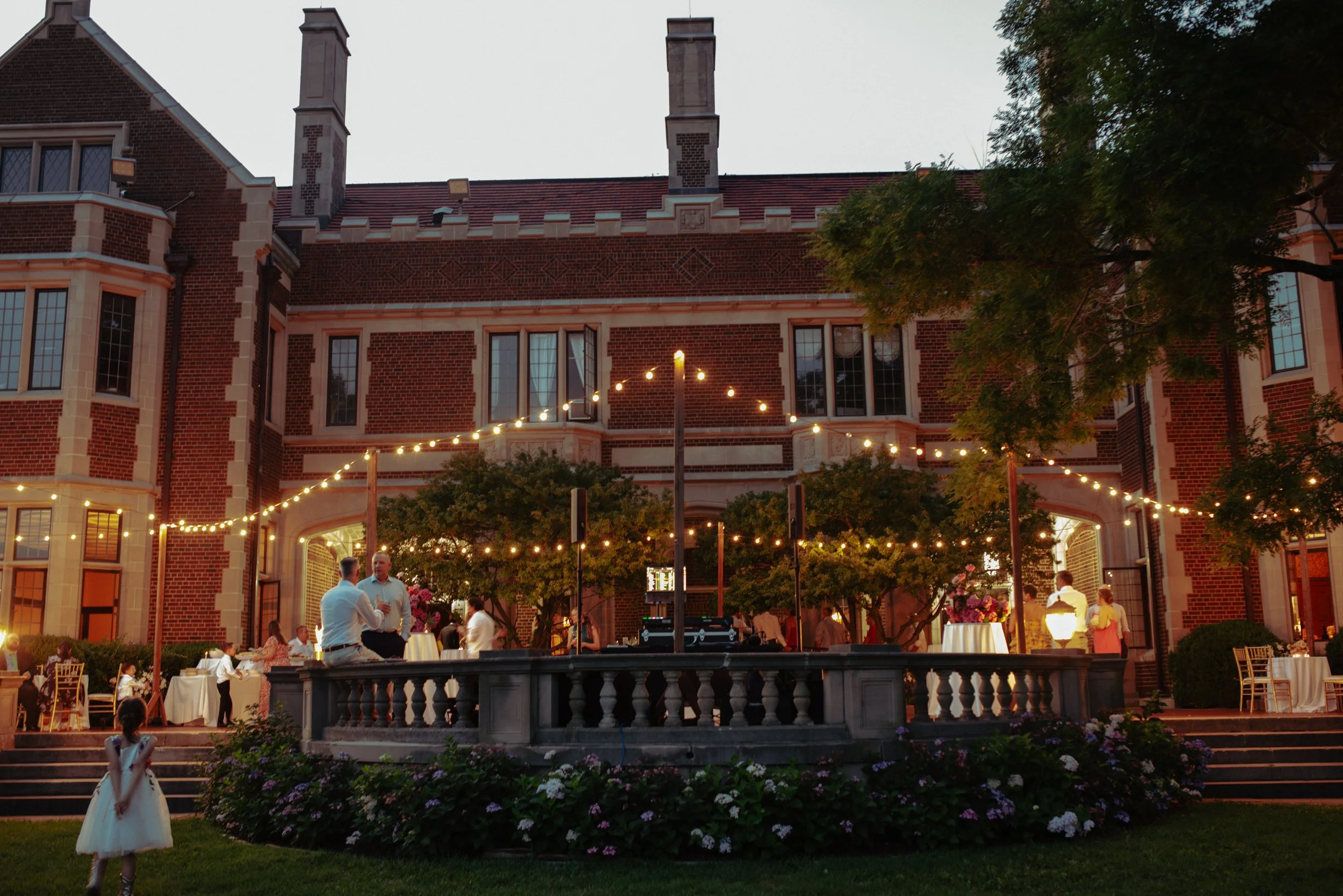 An outdoor evening gathering at a mansion with string lights, people dining at tables, and children playing on the grass, with a large brick building and trees in the background.