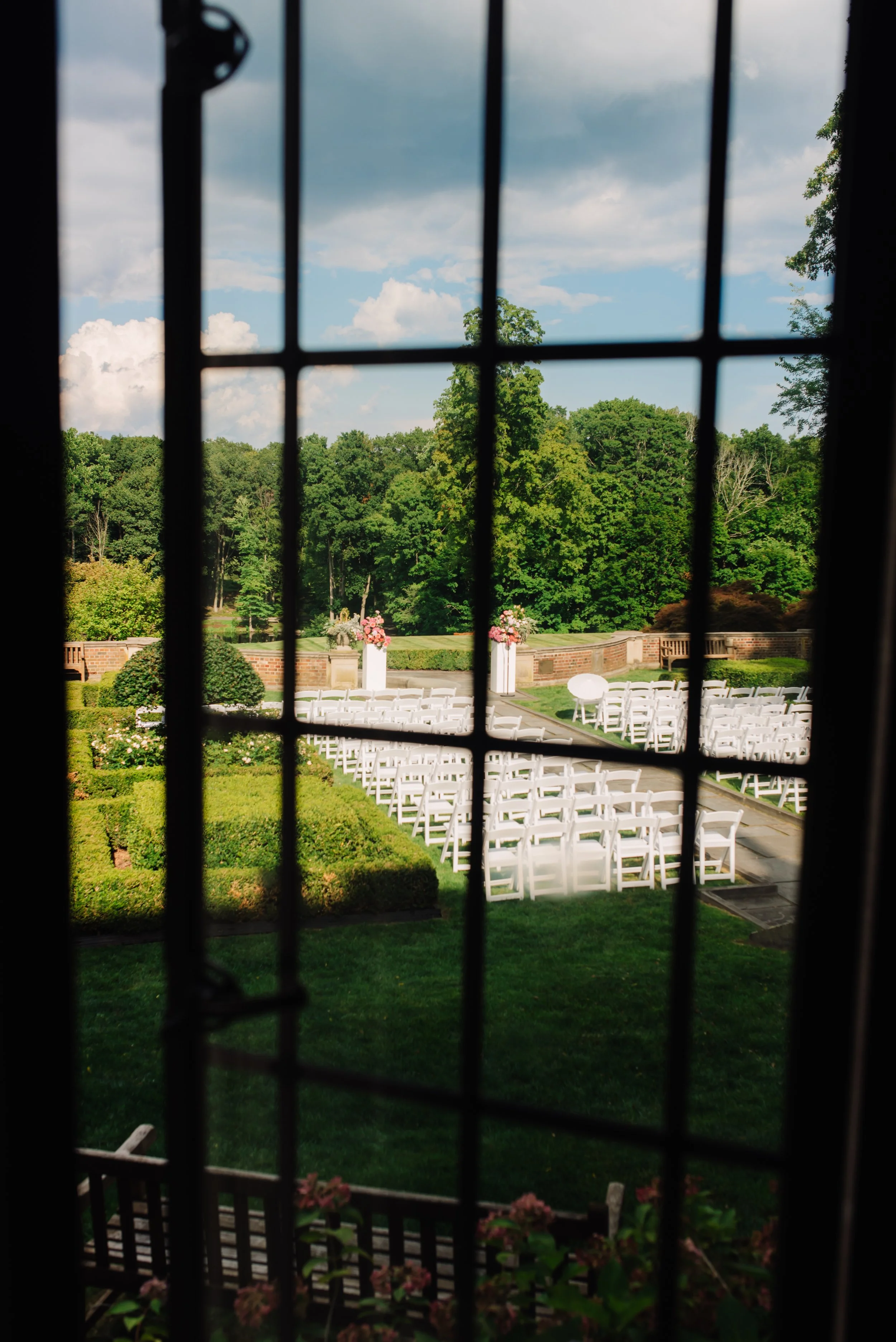 View through a window with black metal grid, showing an outdoor wedding ceremony setup with white chairs arranged in rows, pink floral arrangements on white columns, and a lush green landscape with trees in the background.