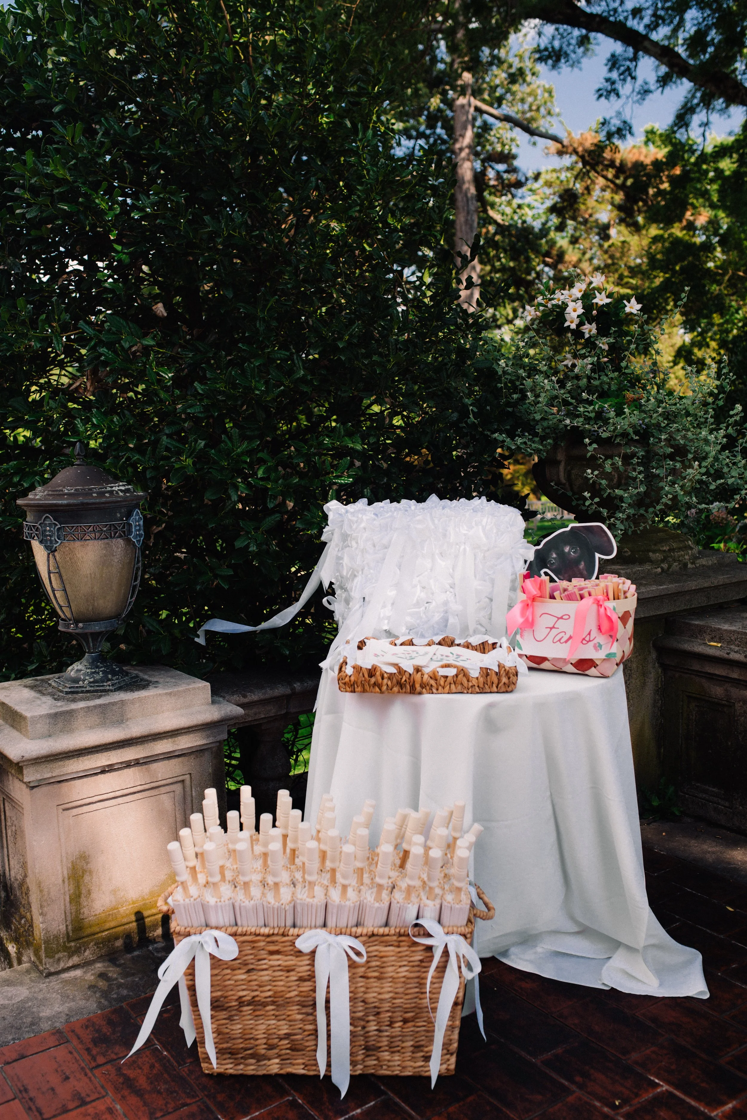 Outdoor dessert table with white cake, striped cookies, and pink wrapped candies, decorated with a basket of white candies with ribbons, surrounded by greenery and a vintage lantern, on a stone and brick patio.