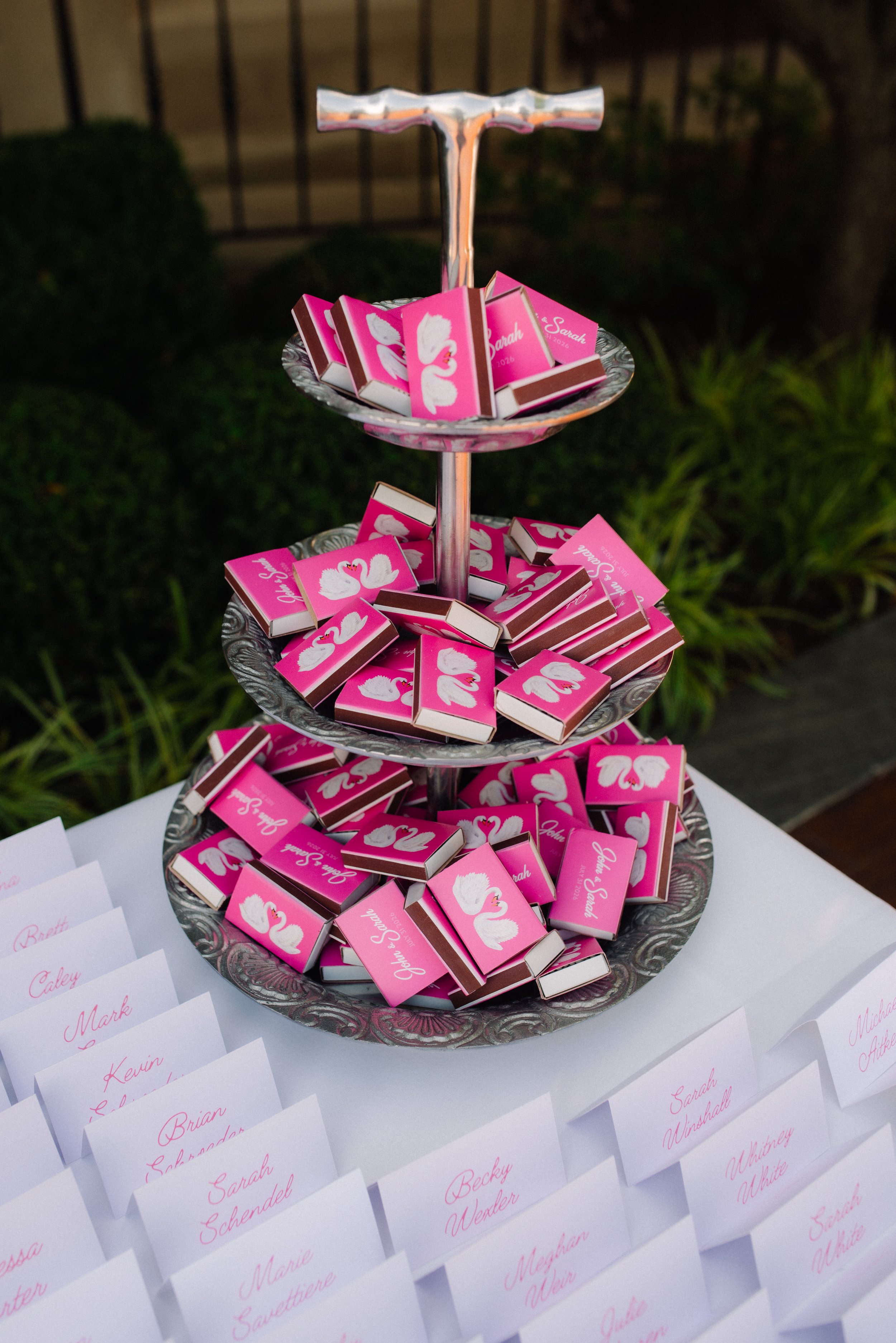 A three-tiered silver tray holding small pink boxes with white swan illustrations, used as place cards at an event, with name cards placed in front on a white tablecloth.