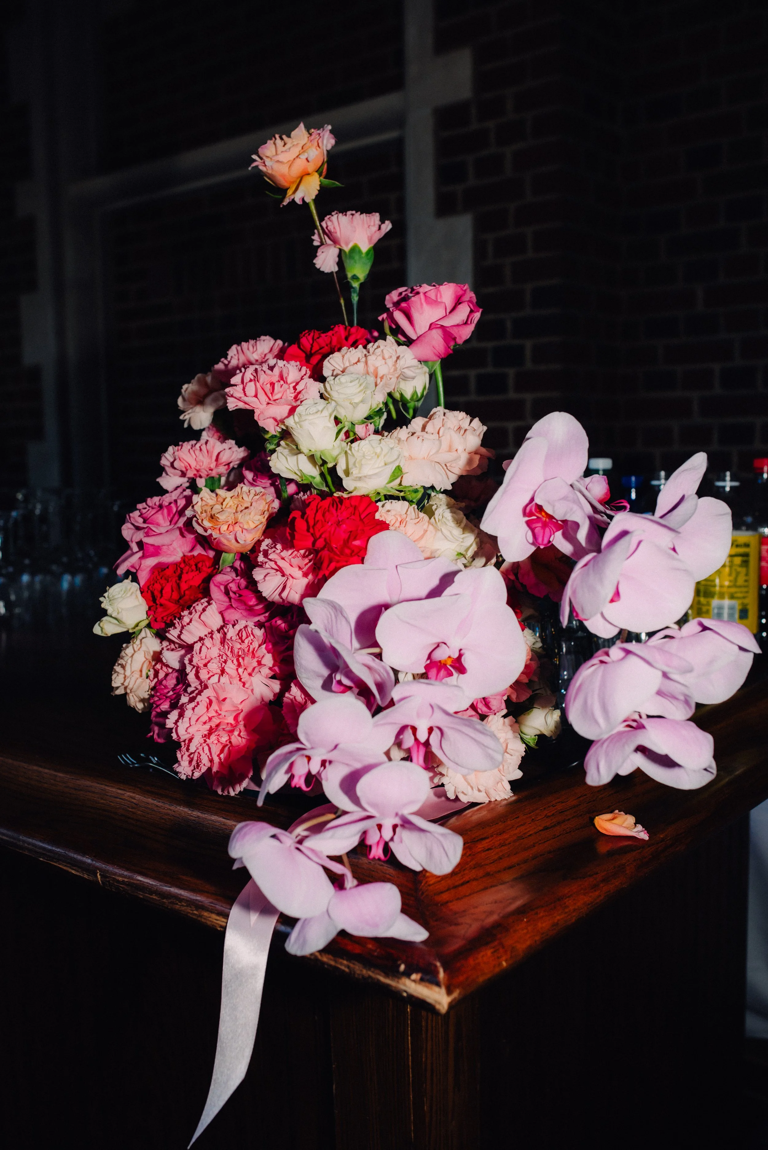 A bouquet of pink and white carnations and orchids arranged on a dark wooden surface with a brick wall in the background.