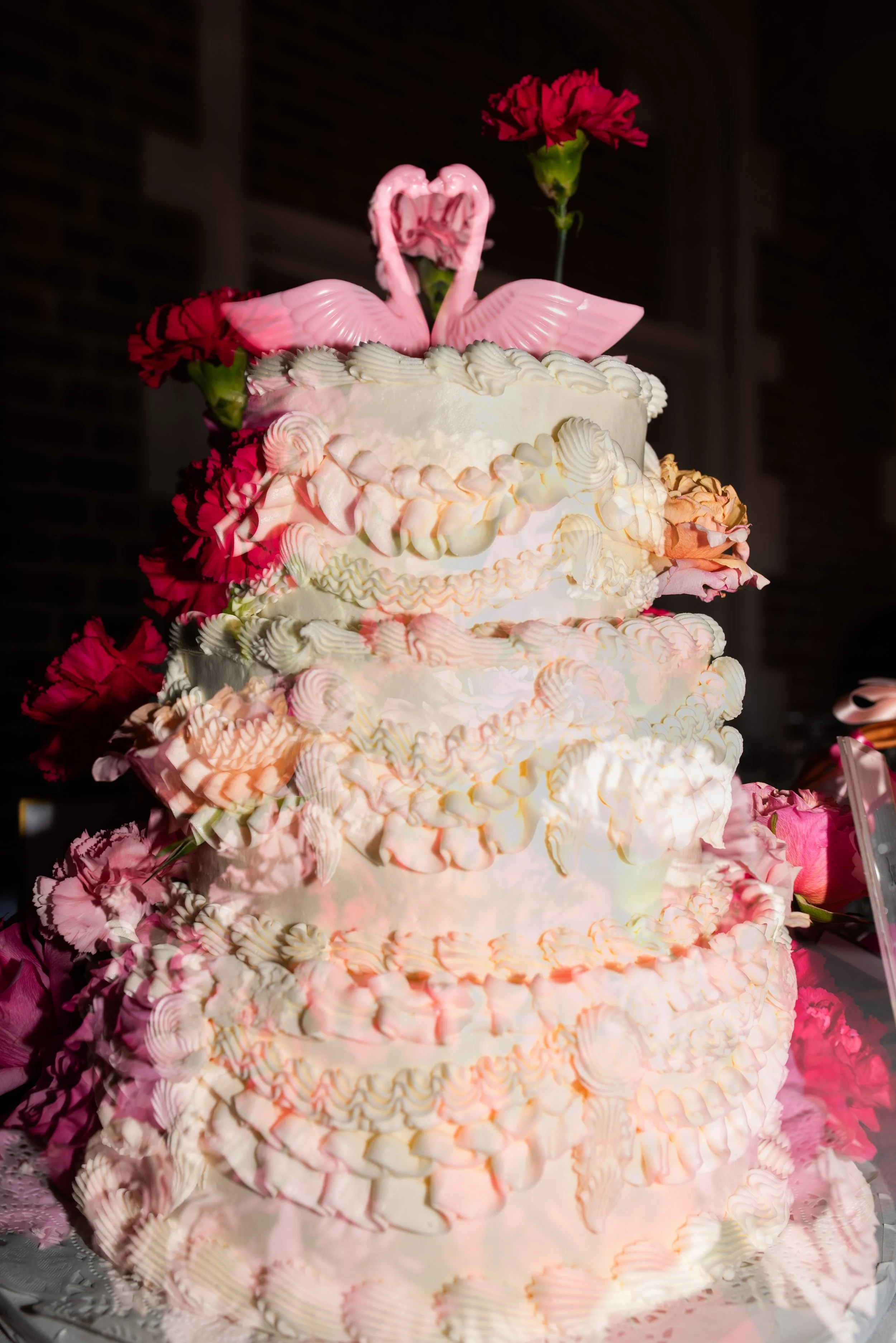 A multi-tiered white wedding cake decorated with pink and white whipped cream swirls and roses, topped with pink swan figurines and pink carnations.