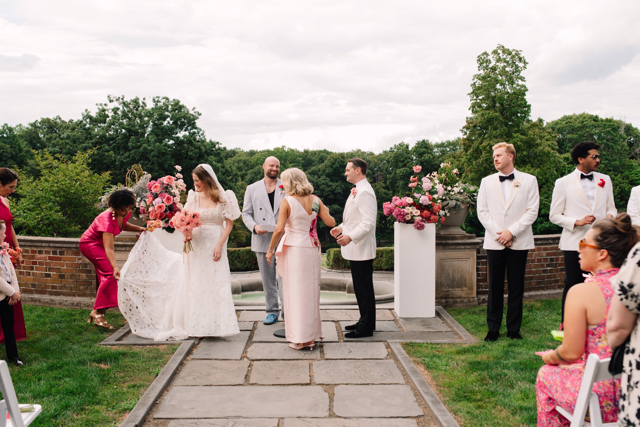 A wedding ceremony outdoors with people dressed in formal attire, holding flowers, with a fountain and trees in the background.