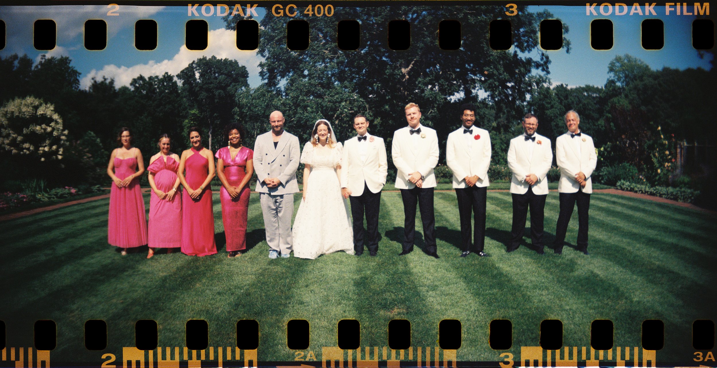 A group of eleven people dressed in wedding attire standing on grass in a park with trees and bushes in the background. The bride is in a white wedding gown, and the groom and other men are in white tuxedos with black bow ties. The women on the left are in pink dresses.