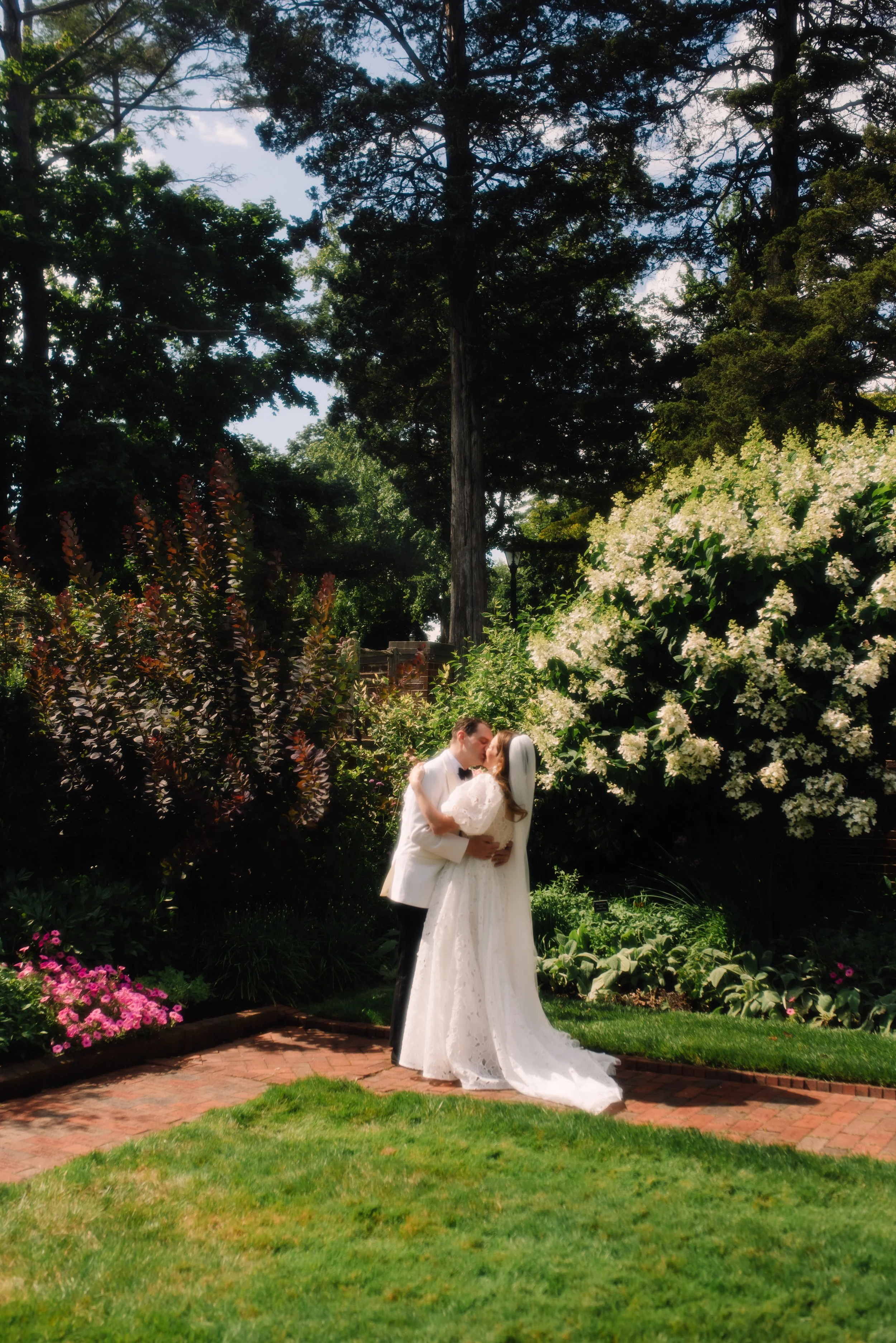 A bride and groom in wedding attire sharing a kiss in an outdoor garden surrounded by trees, flowers, and greenery.