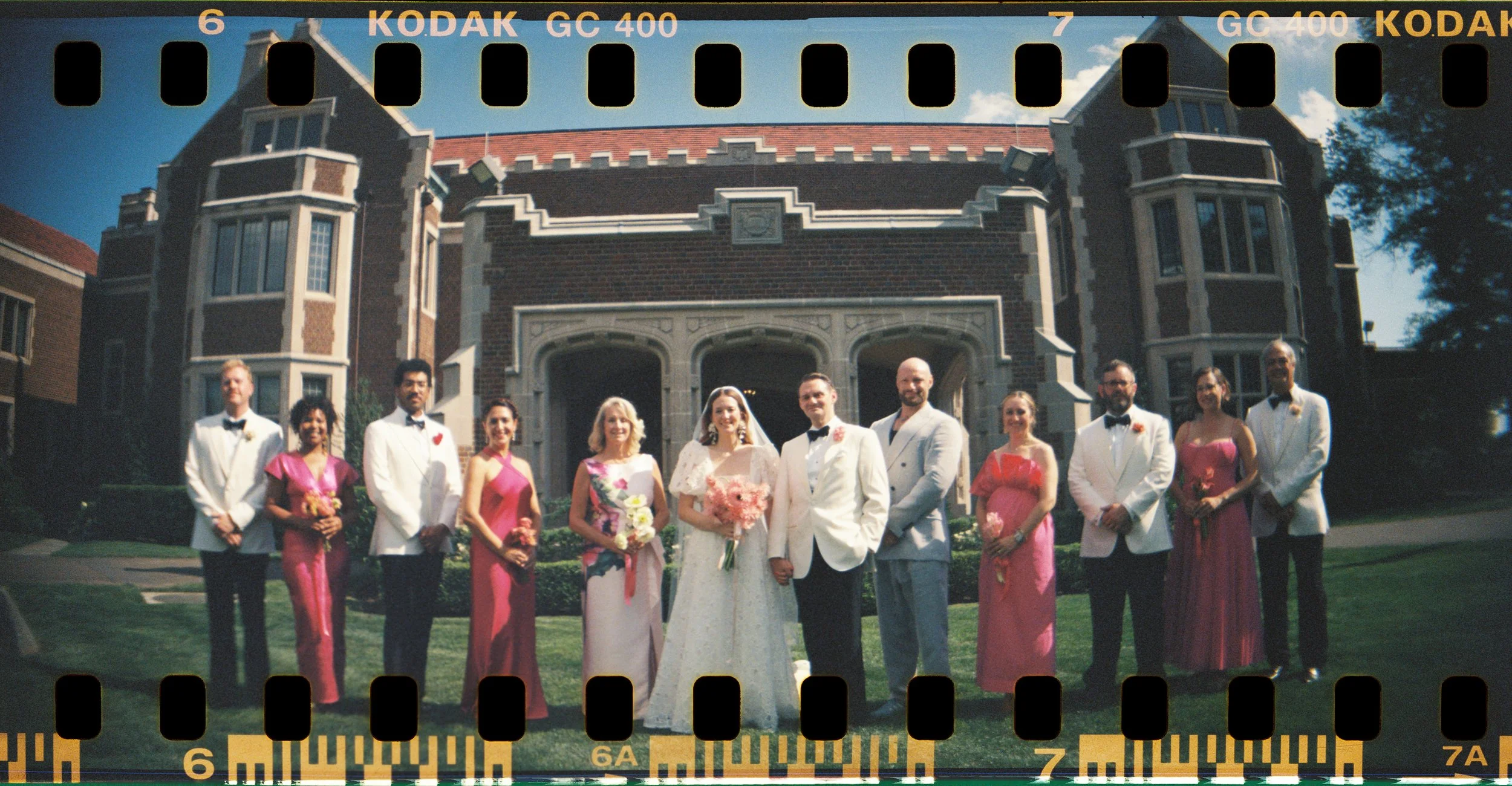 A group of ten people in formal wedding attire standing outside in front of a large brick house with turrets and a front porch, under a blue sky. The group includes men and women, with some women in pink dresses and the bride in a white wedding gown holding a bouquet of pink flowers.
