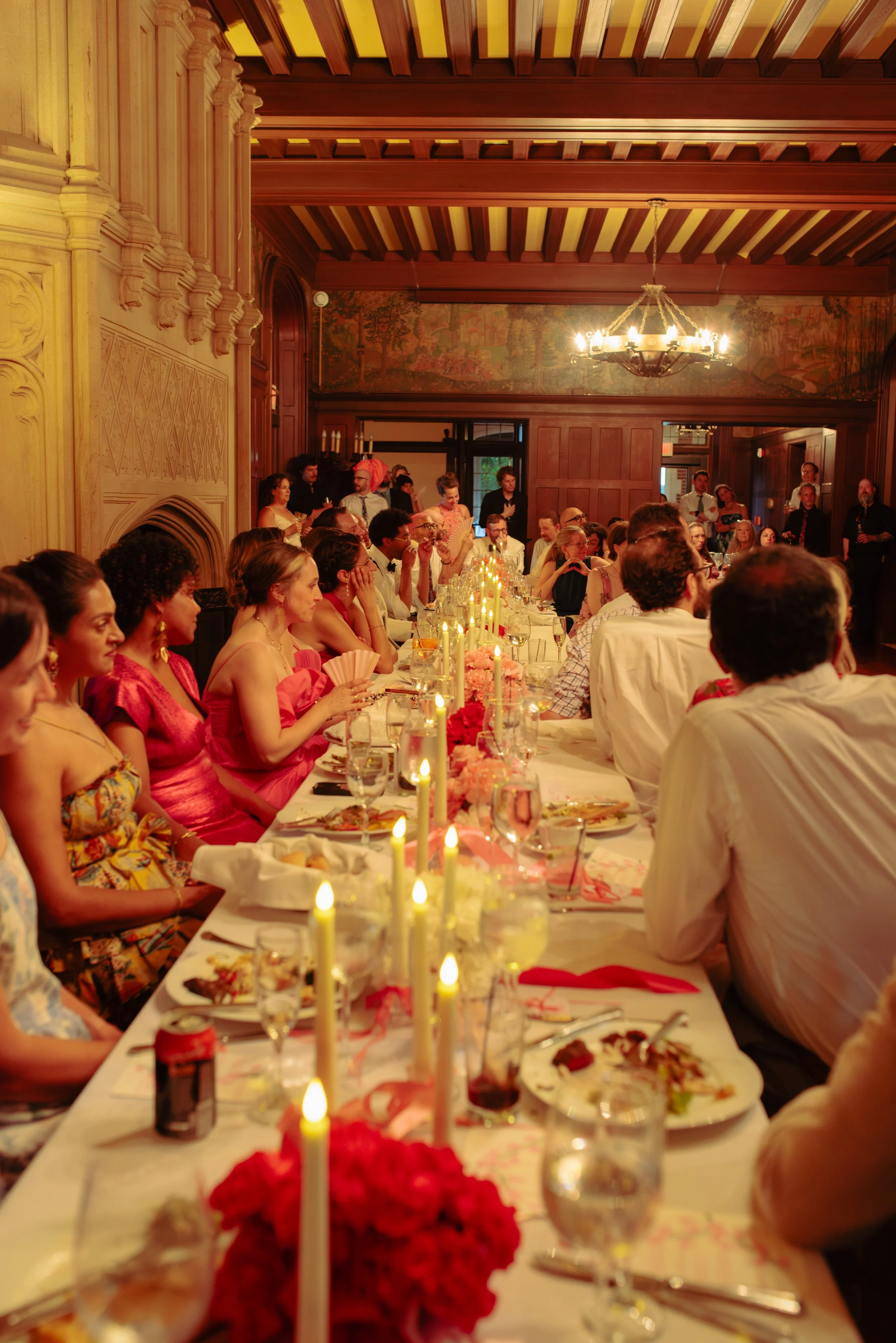 A large group of people seated at a decorated dinner table in a wood-paneled room with a chandelier, candles, and a mural on the wall, during a formal celebration or event.