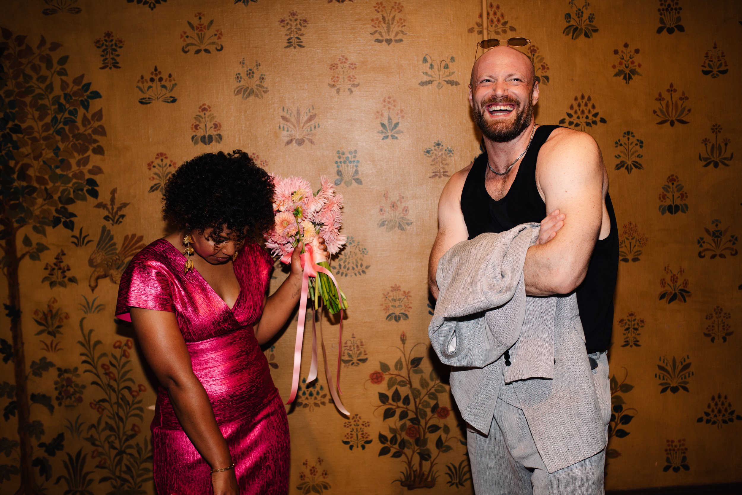 A woman in a pink dress holding pink flowers and a man smiling with sunglasses on his head and a gray suit jacket draped over his arm, standing in front of a floral wallpaper background.