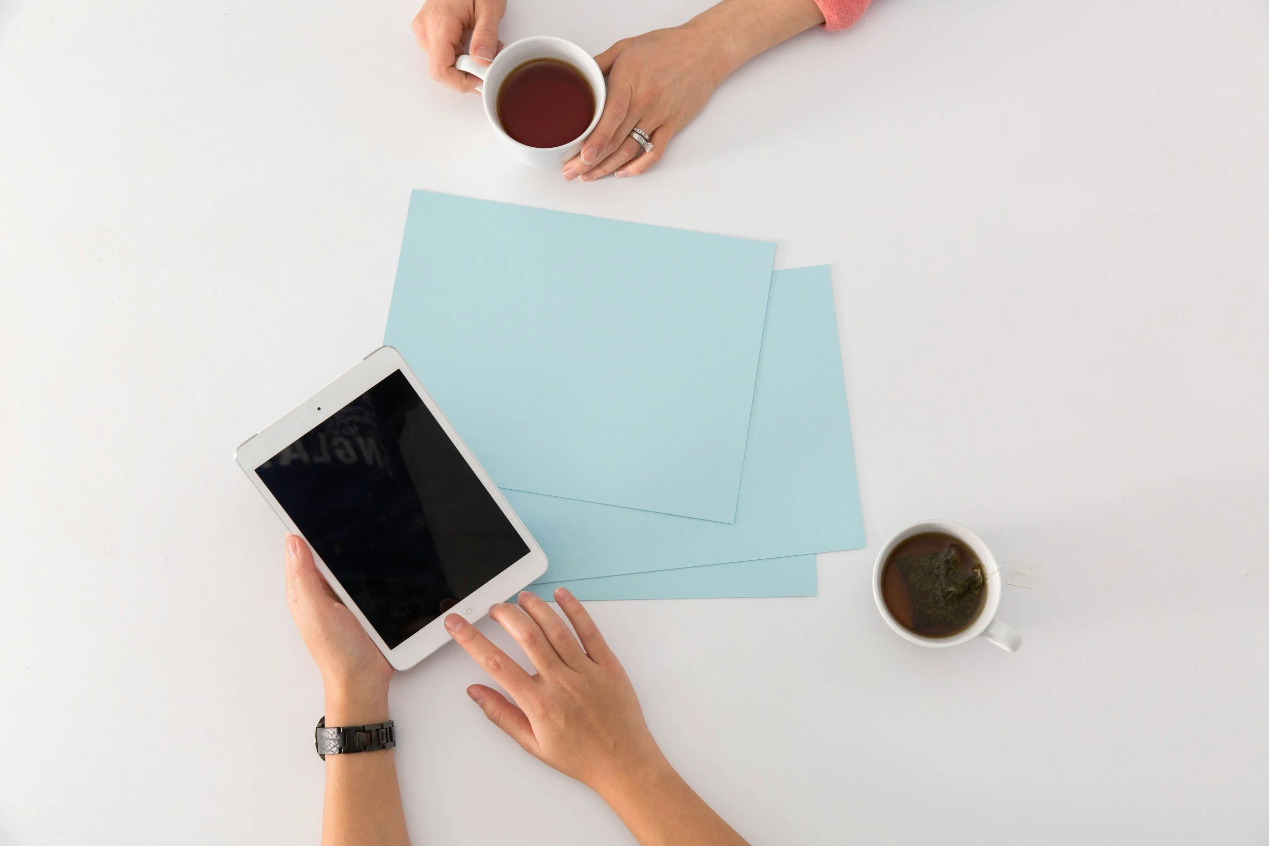 Overhead view of a person holding a white smartphone and a cup of tea, with another cup of tea and two blue papers on a white table.
