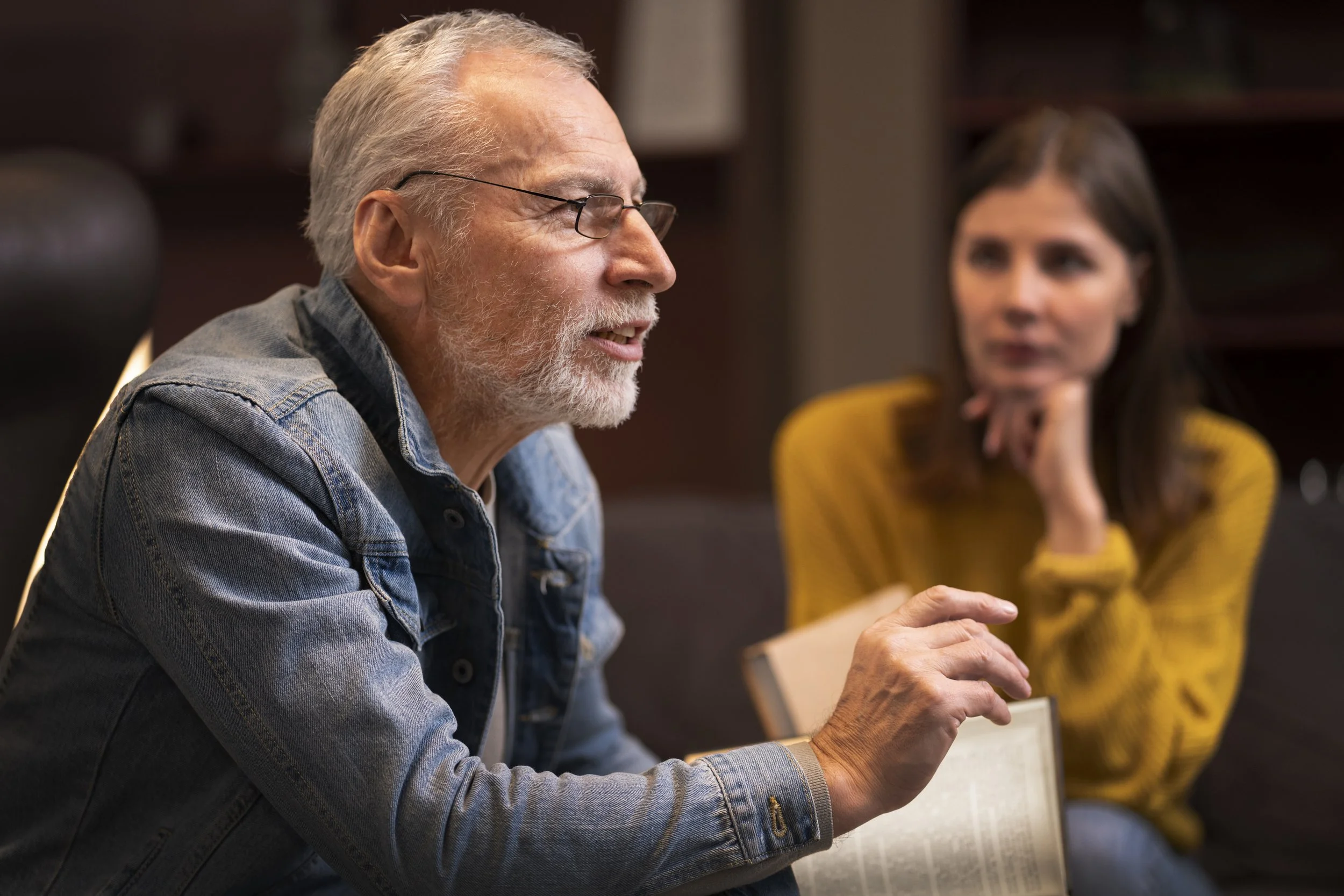 An elderly man with gray hair, glasses, and a beard talking, with a woman in a yellow sweater listening attentively.
