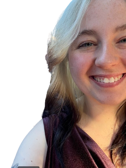 A young woman smiling with blonde hair and blue eyes, wearing a burgundy top, taking a selfie outdoors.