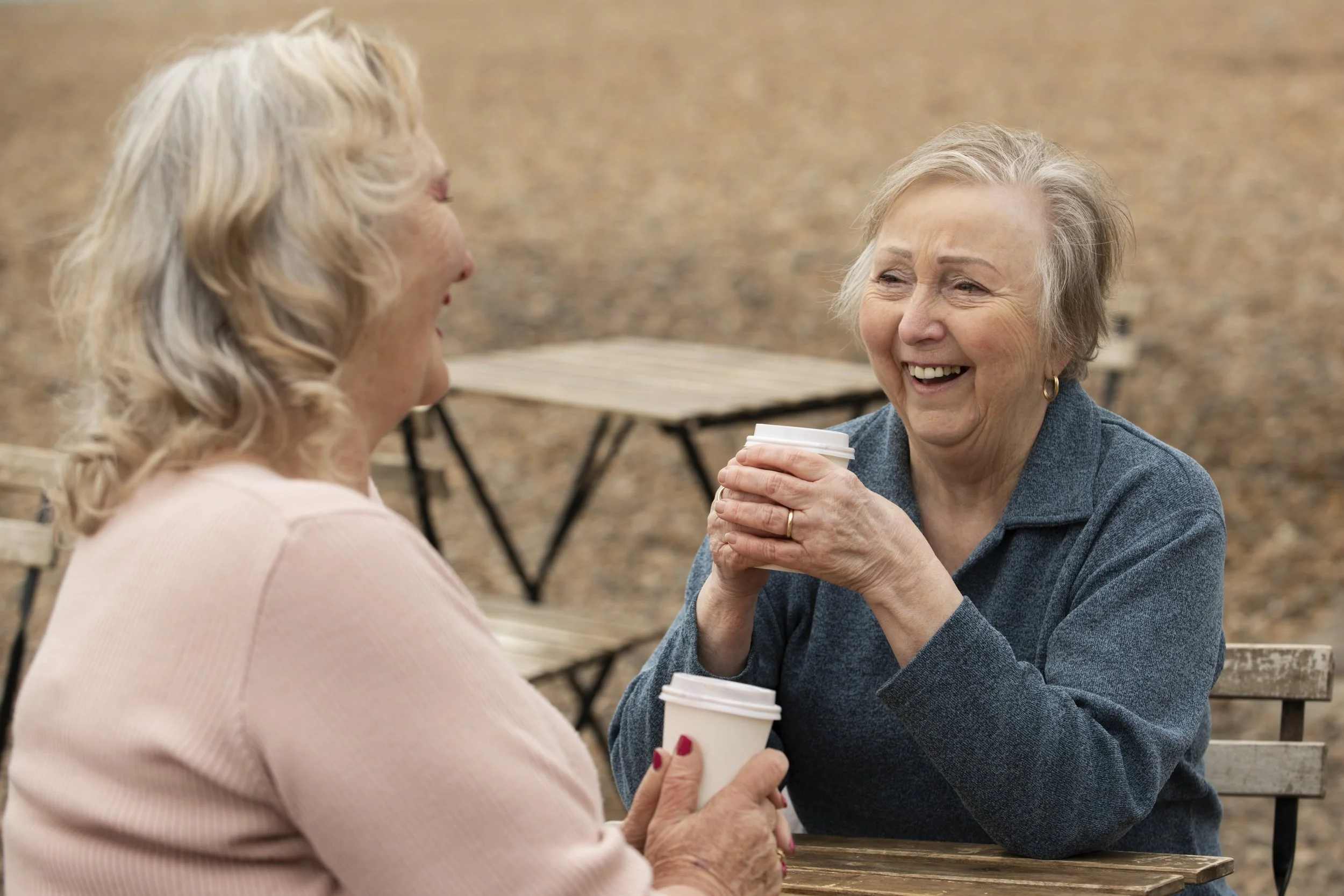 Two elderly women laughing and talking while holding coffee cups outdoors on a wooden table.
