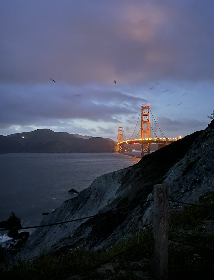 Nighttime view of the Golden Gate Bridge illuminated, with a cloudy sky and mountains in the background, seen from a rocky shore with a wooden fence and grassy foreground.