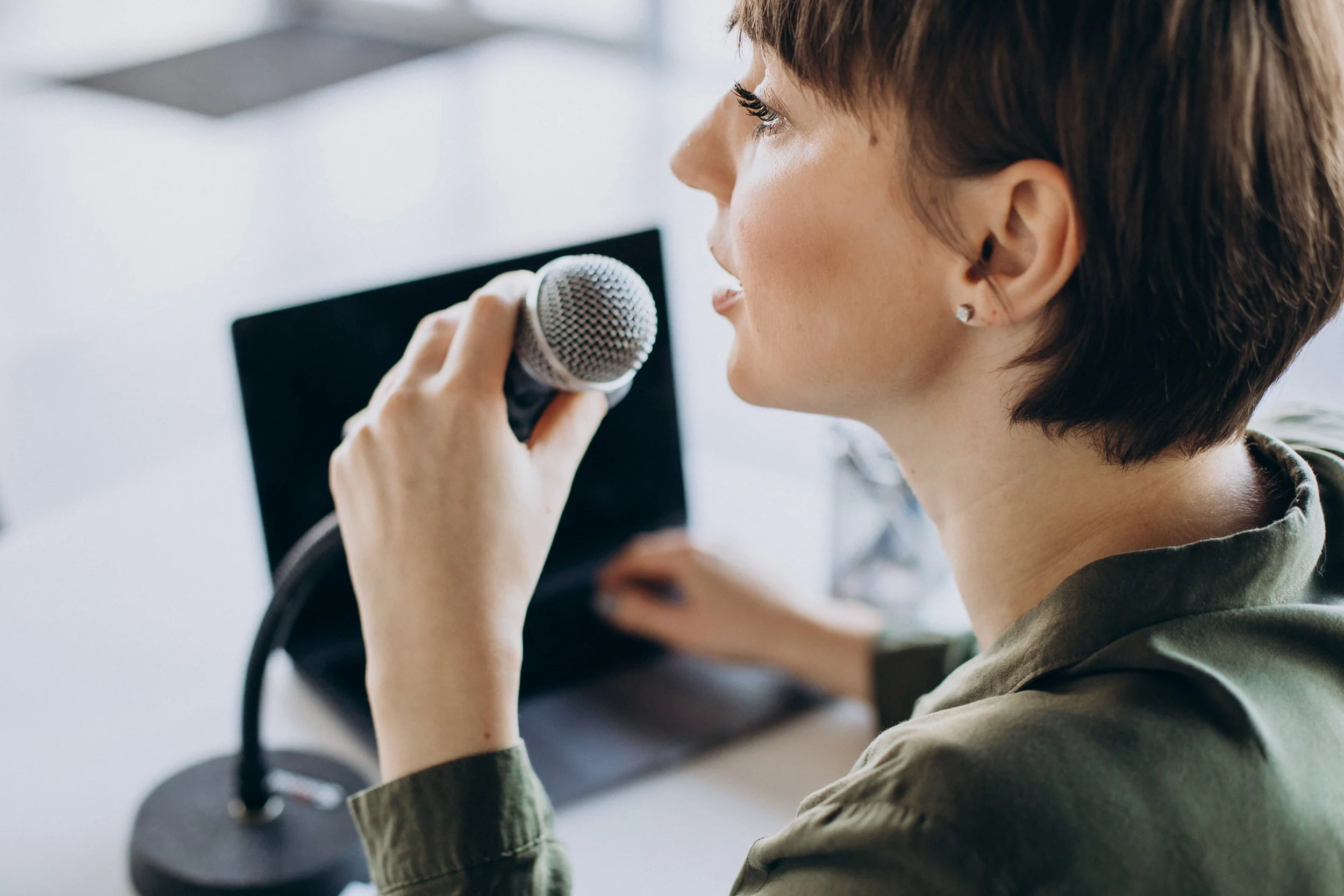 A woman with short brown hair speaking into a microphone near a computer monitor.